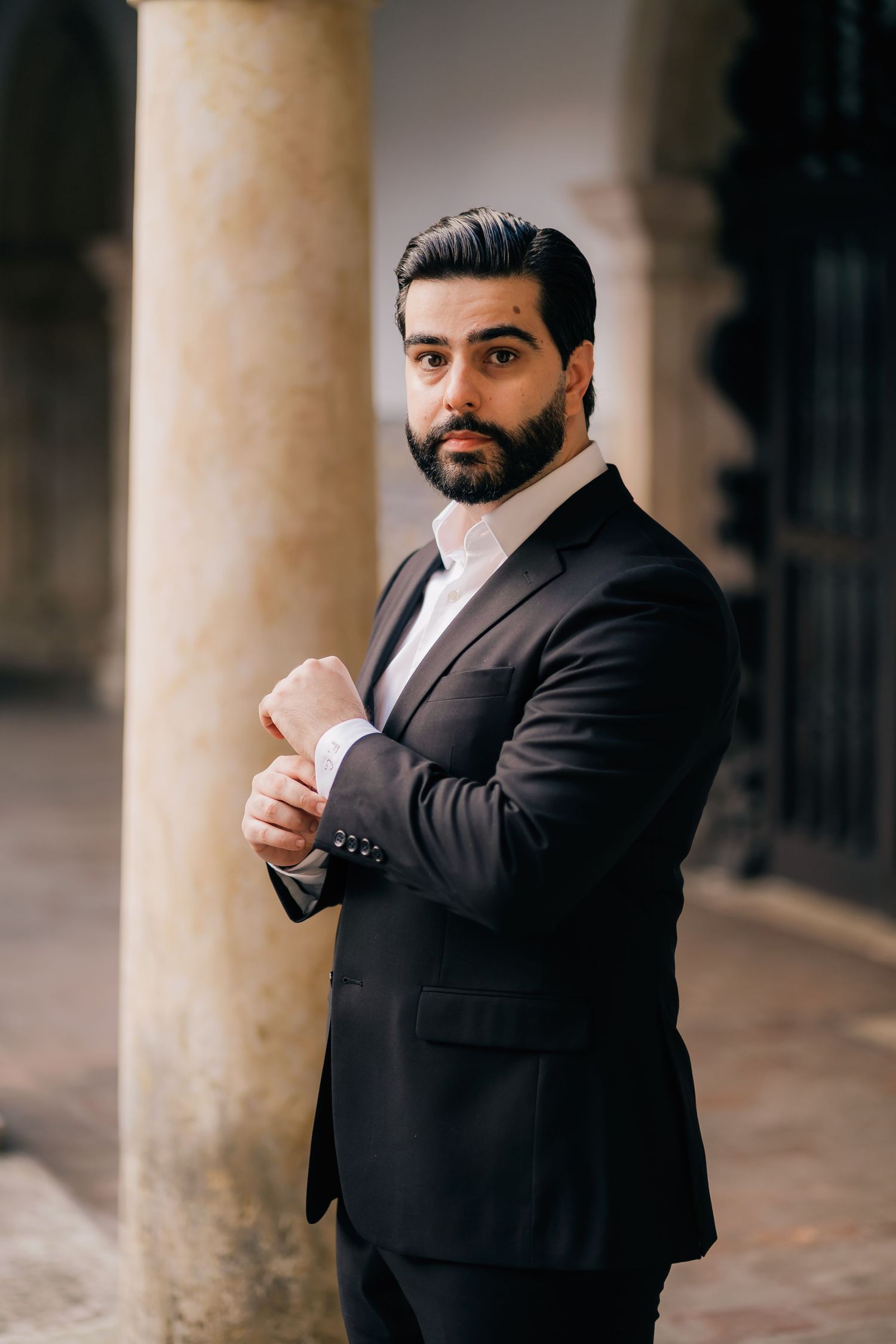 A person in a dark suit poses dramatically against classical columns.
