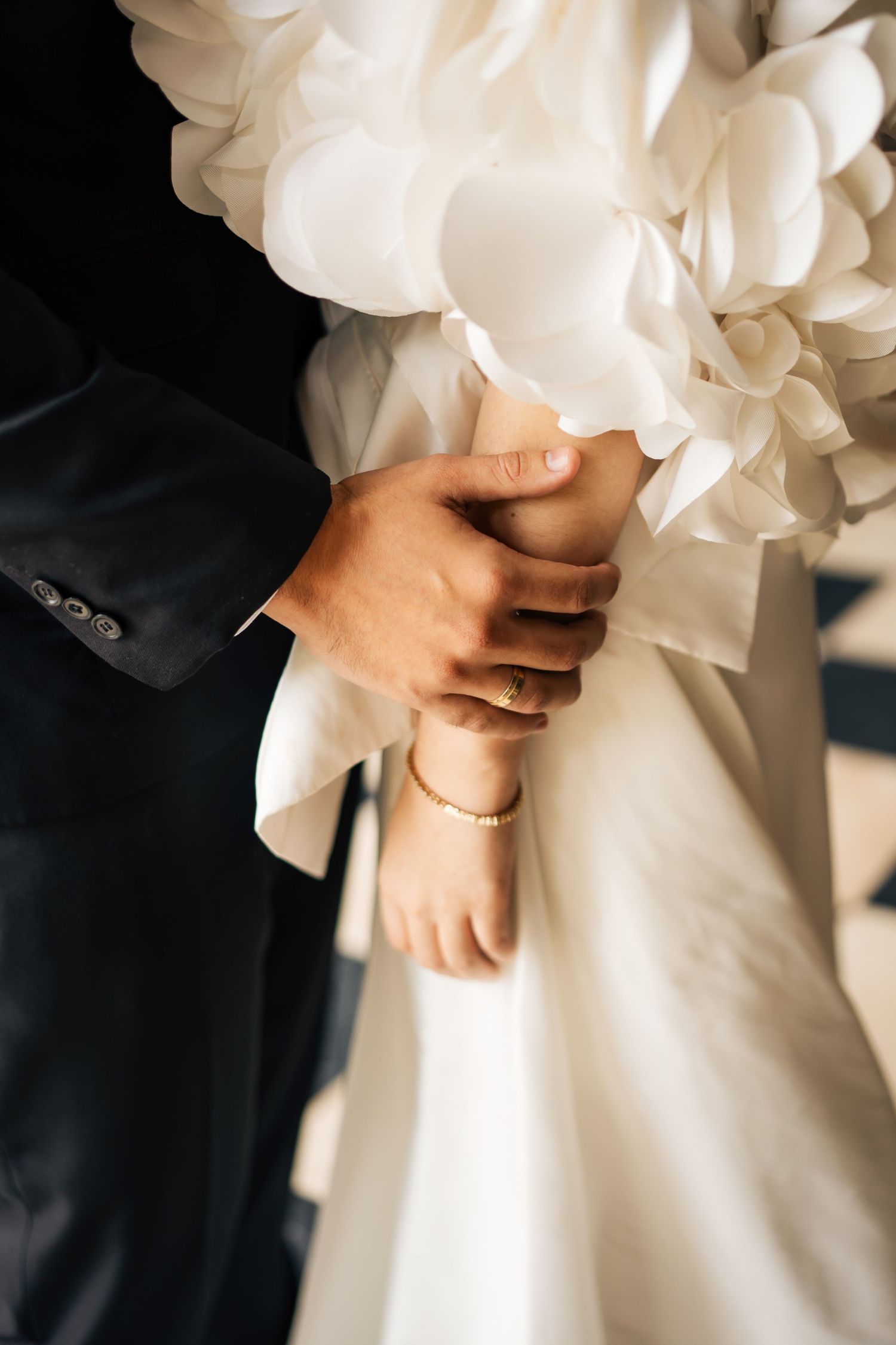 A close-up of hands touching at a wedding, with a black suit sleeve and flowing white bridal dress fabric visible.