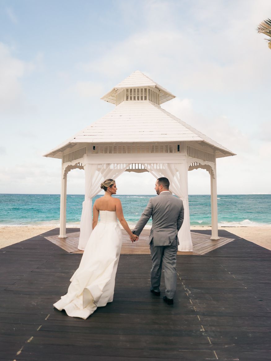 A couple sits together on a wooden pier with a white gazebo overlooking turquoise ocean waters on their wedding day.