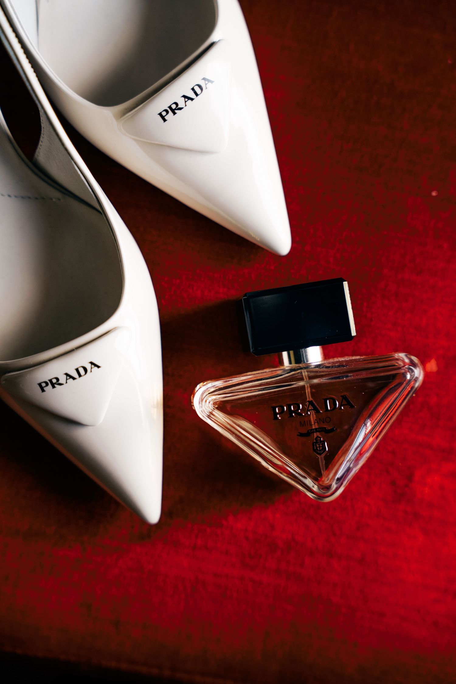 White Prada high heel shoes and perfume bottles arranged on red velvet background in artistic product photography series.