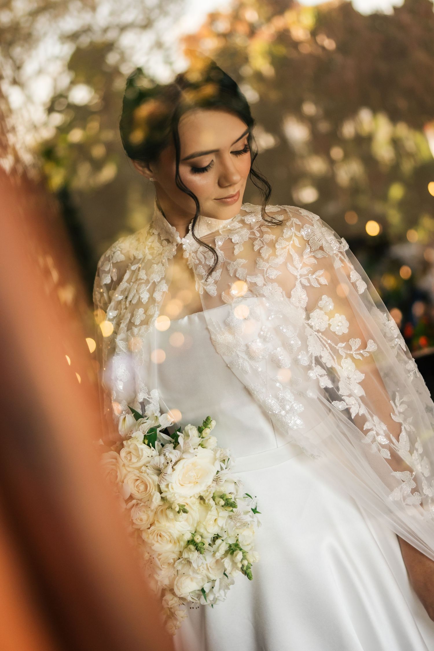 Close-up details of delicate white lace wedding dress with floral embroidery and soft bokeh lighting effect in background.