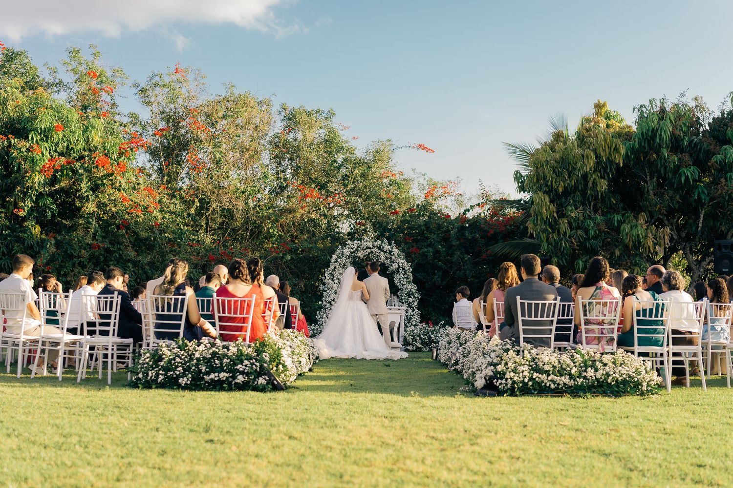 An outdoor garden wedding ceremony setup with white chairs arranged in rows and floral decorations along a grassy aisle.
