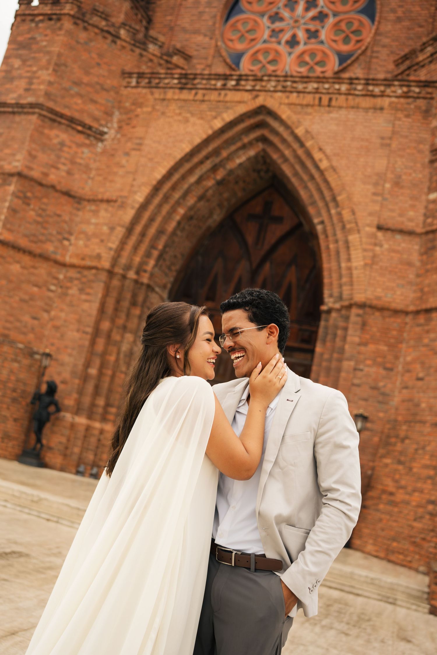 A couple shares an intimate moment in front of a historic brick church with Gothic arches.
