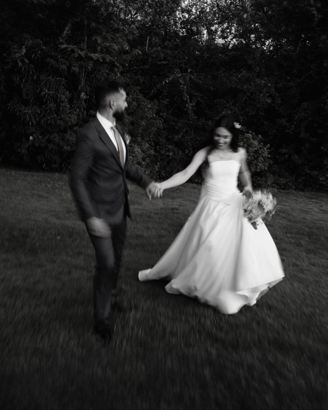 A couple in wedding attire dances and twirls together on a grassy outdoor area at dusk.
