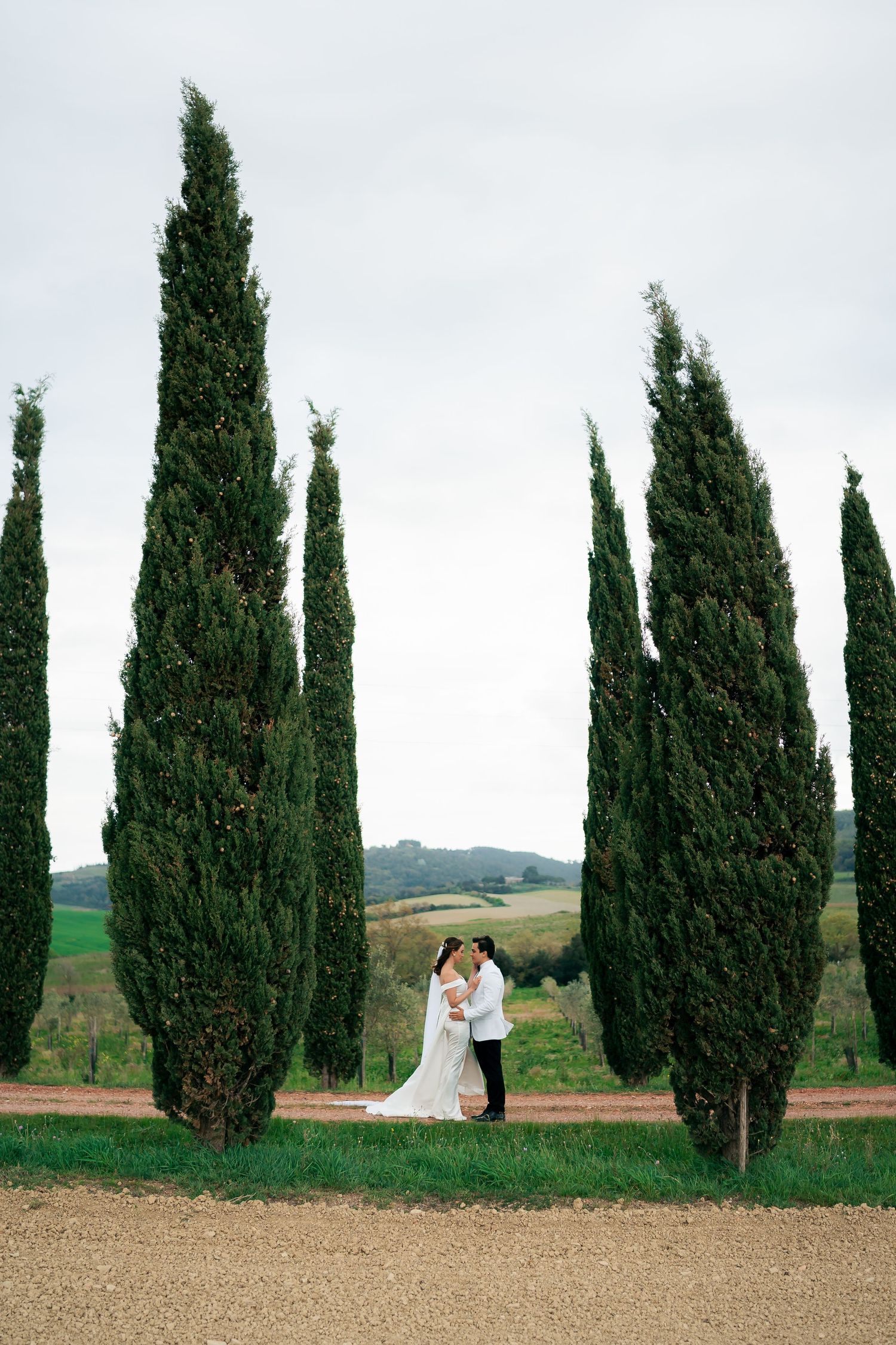 Bride and groom embracing among tall cypress trees in Tuscany, with rolling green hills in the background.