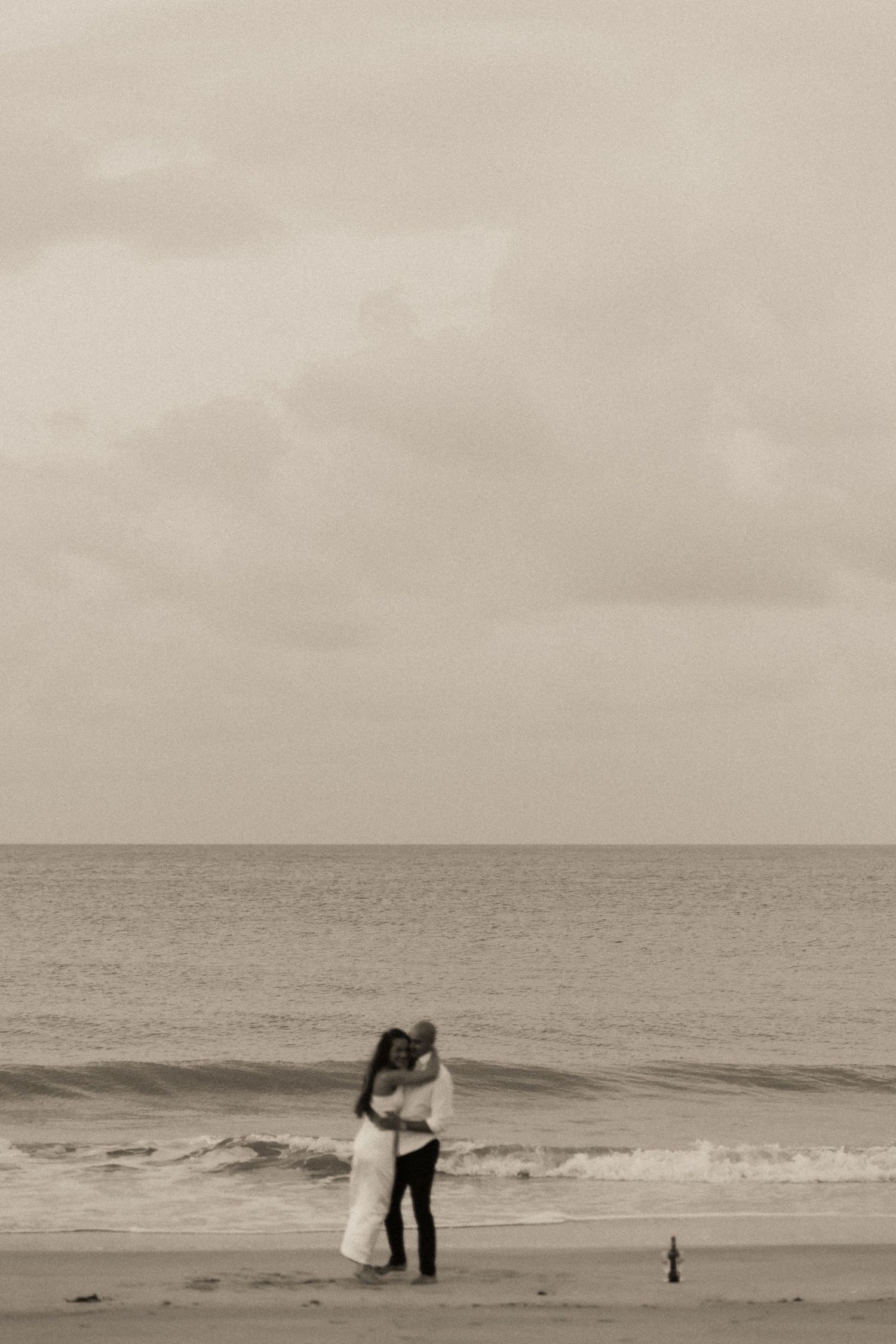 A black and white panoramic sequence shows figures walking and embracing on a misty beach with ocean waves.