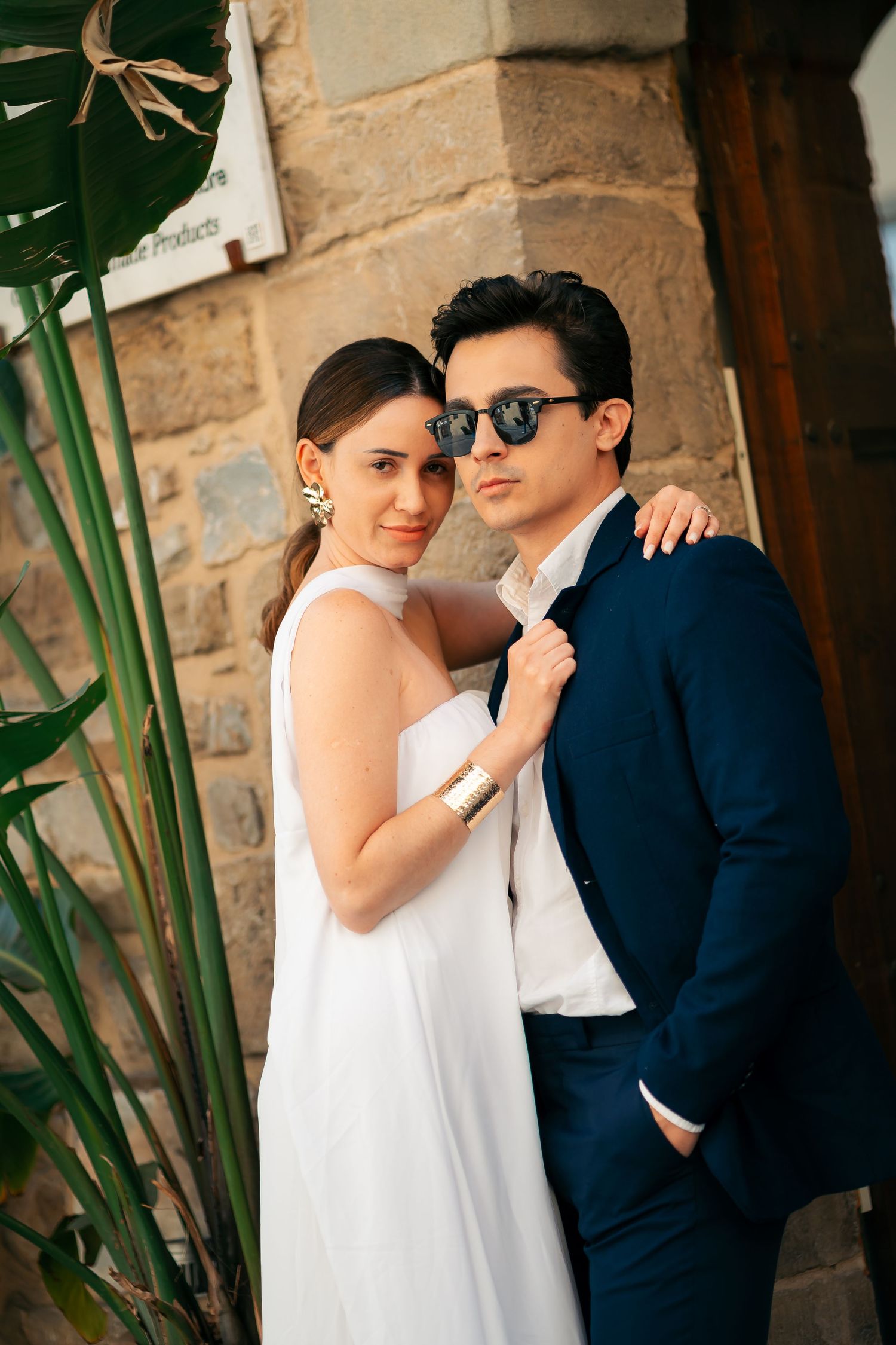A couple in wedding attire pose against a stone wall with greenery, wearing a white dress and navy suit.