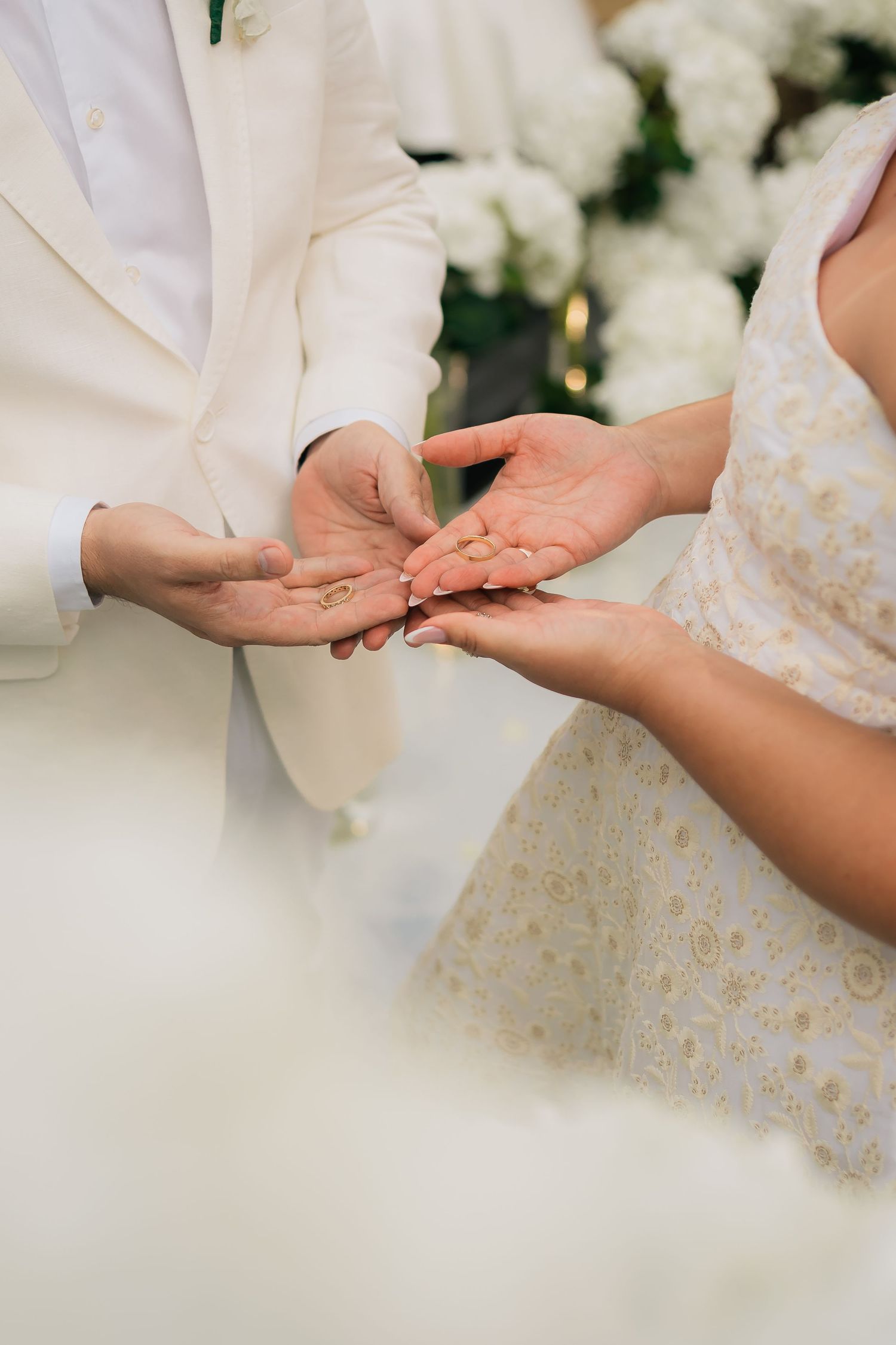 Close-up view of hands exchanging wedding rings during a ceremony with white floral decorations in the background.