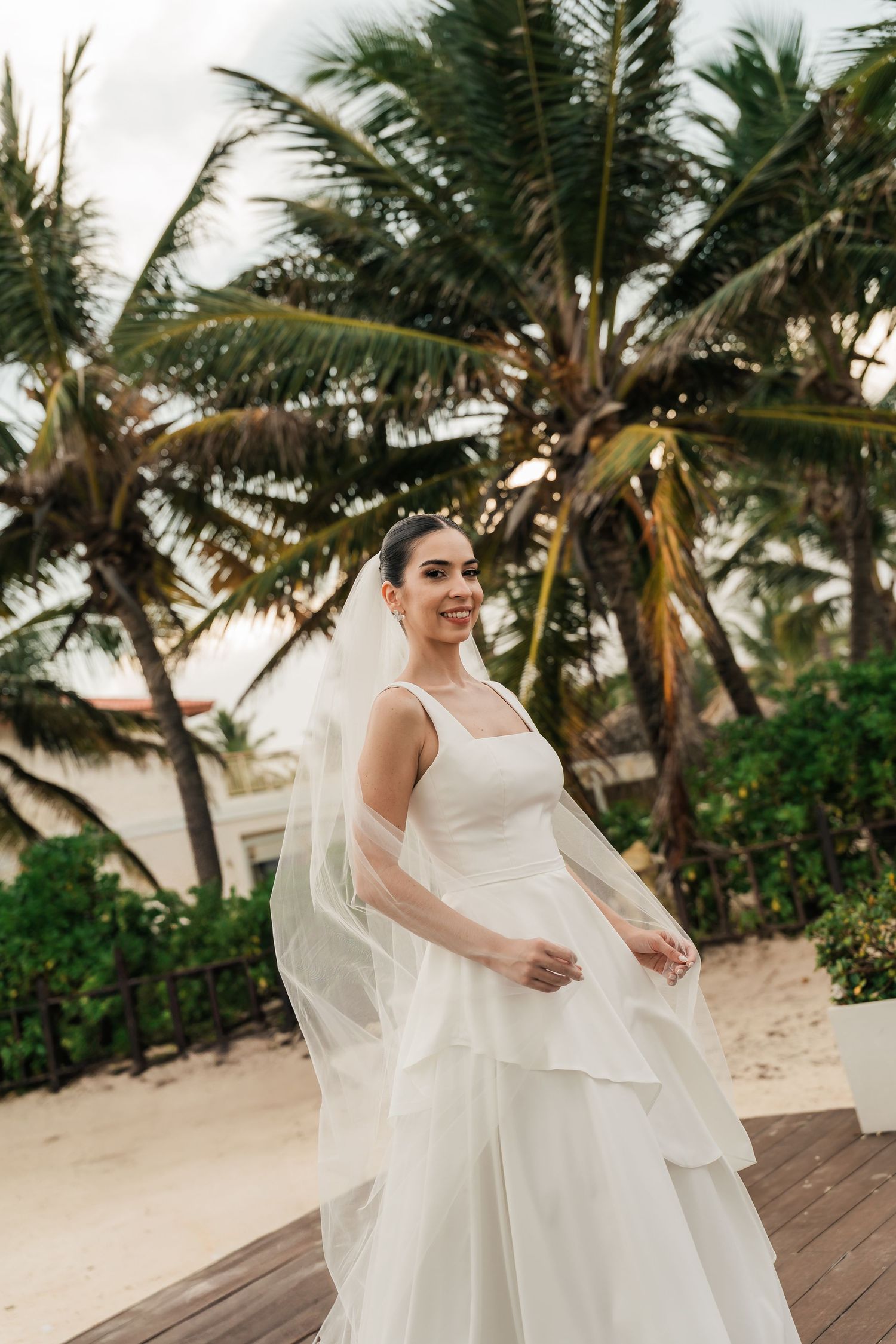 A flowing white wedding dress billows in the tropical breeze among palm trees on a beach deck.