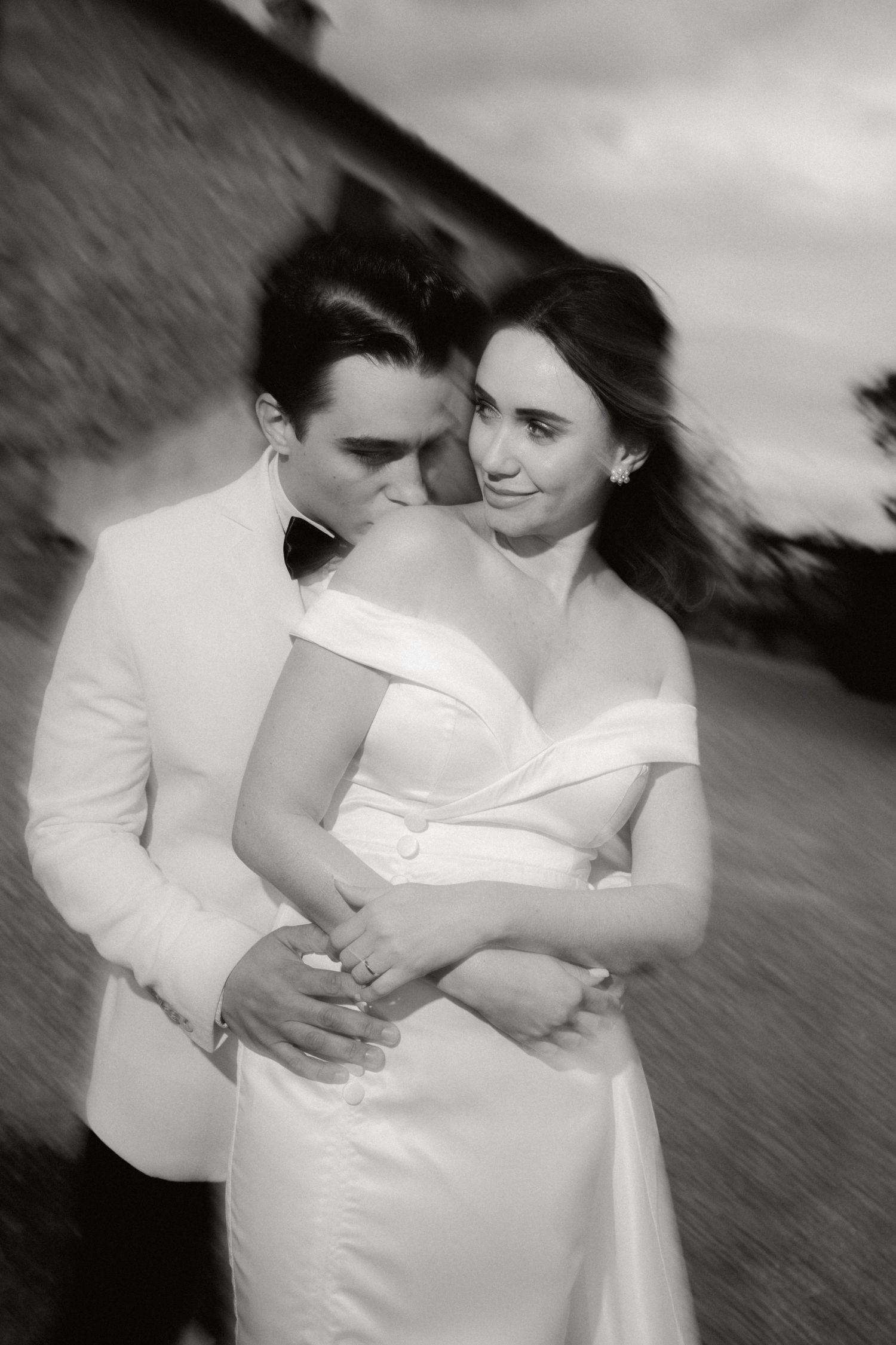 A romantic black and white photo of a groom in a white tuxedo embracing his bride in an off-shoulder gown.
