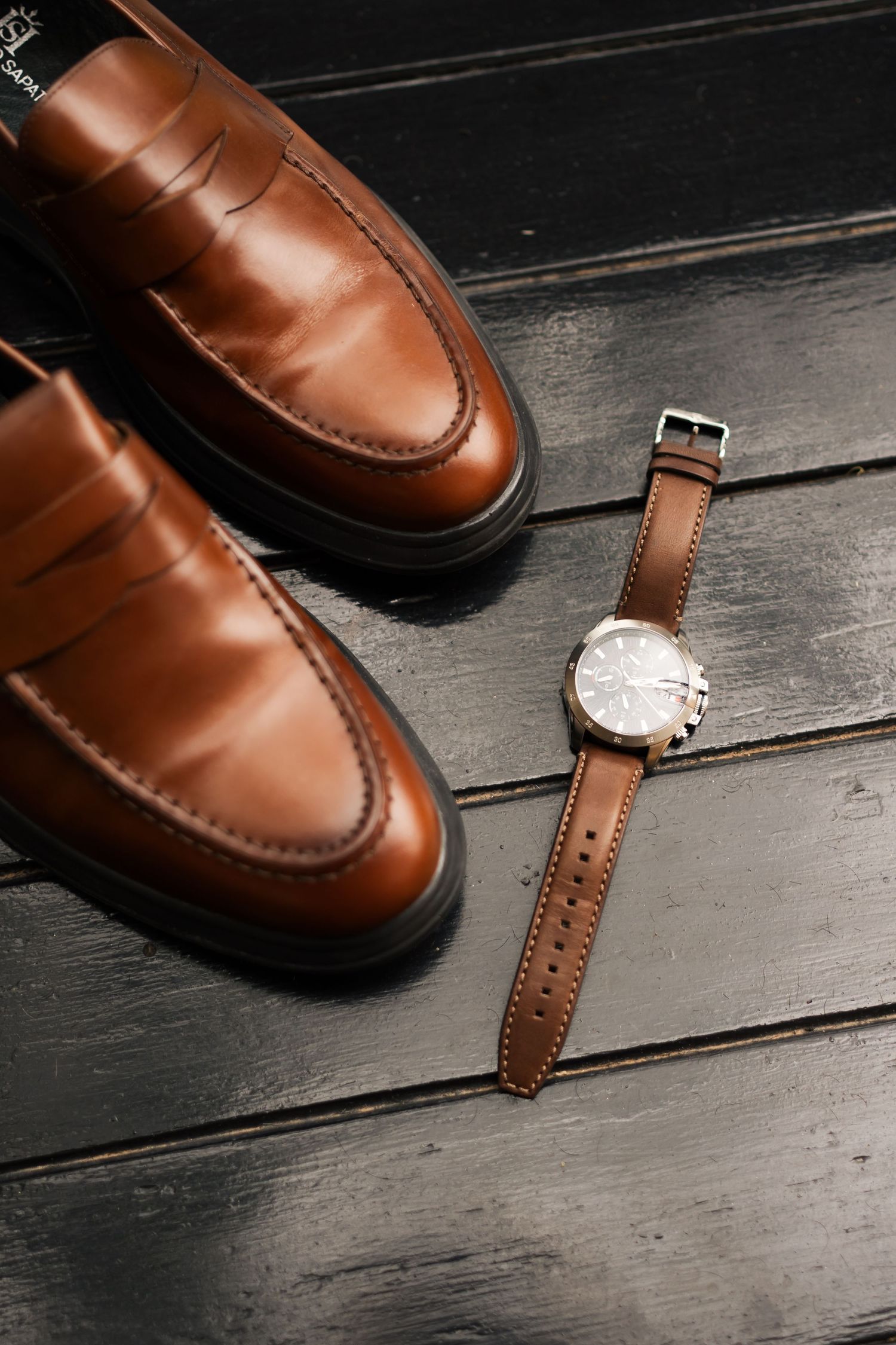 Brown leather dress shoes and matching leather watch strap displayed on dark wooden flooring.