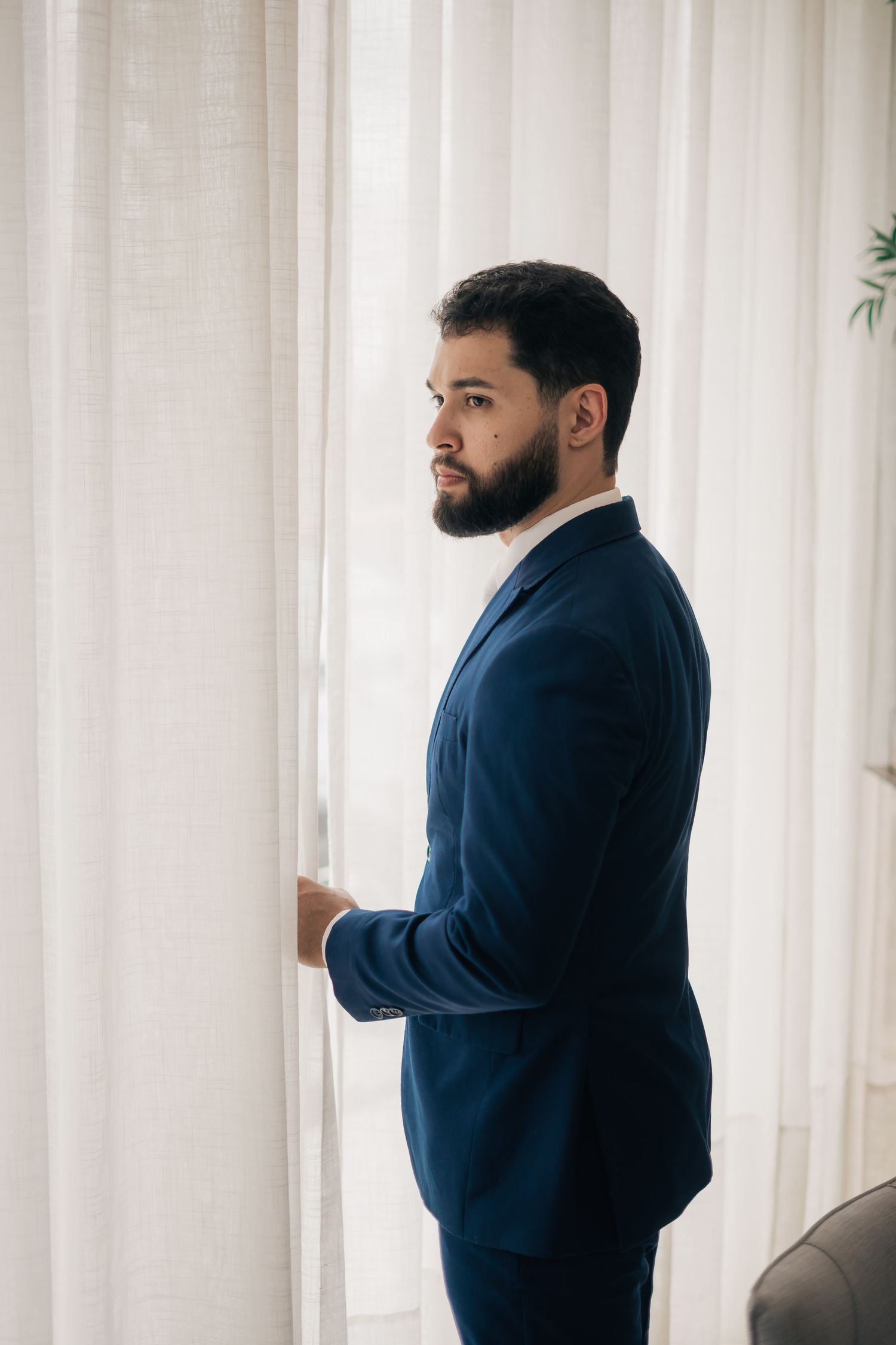 A person in a navy suit looks out through white curtains in an elegant room.