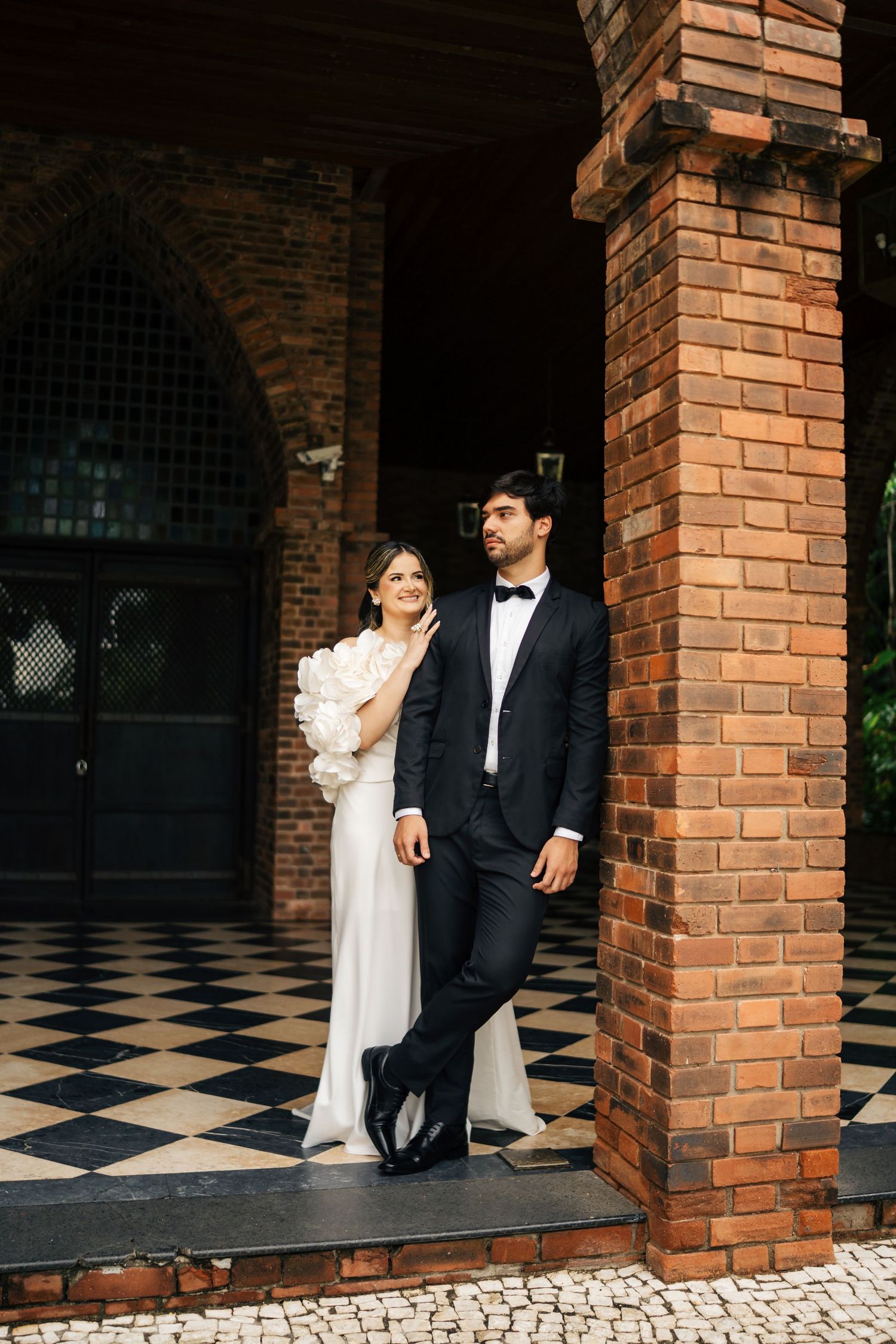 Romantic couple in formal attire poses together against brick columns with black and white checkered floor.