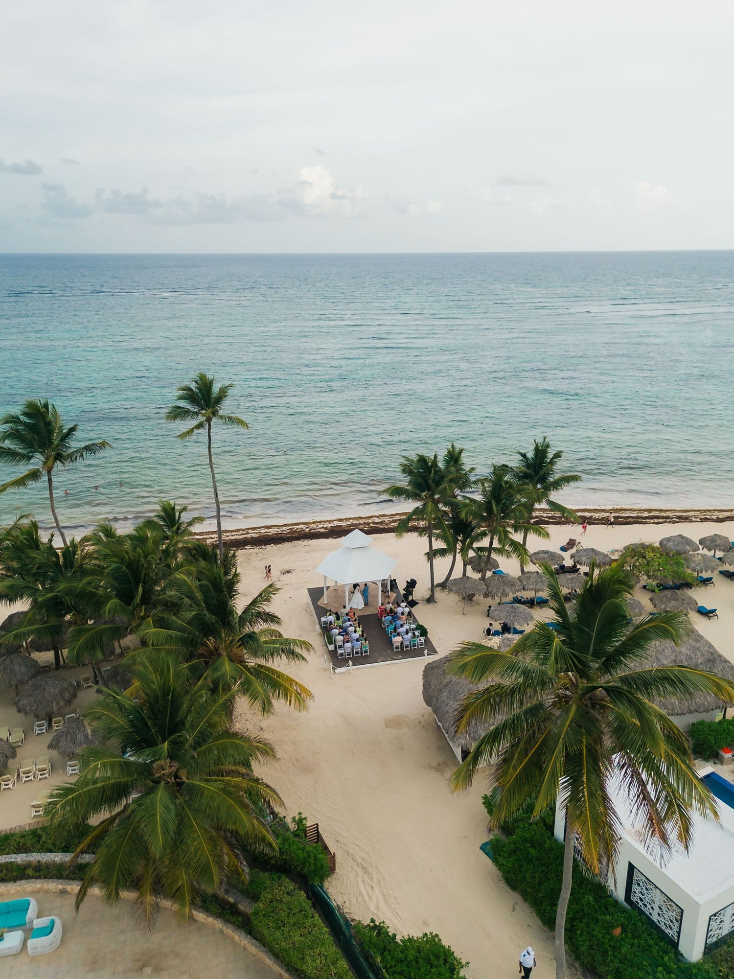 Aerial view of a tropical beach resort with palm trees, thatched umbrellas, lounge chairs, and turquoise ocean waters.