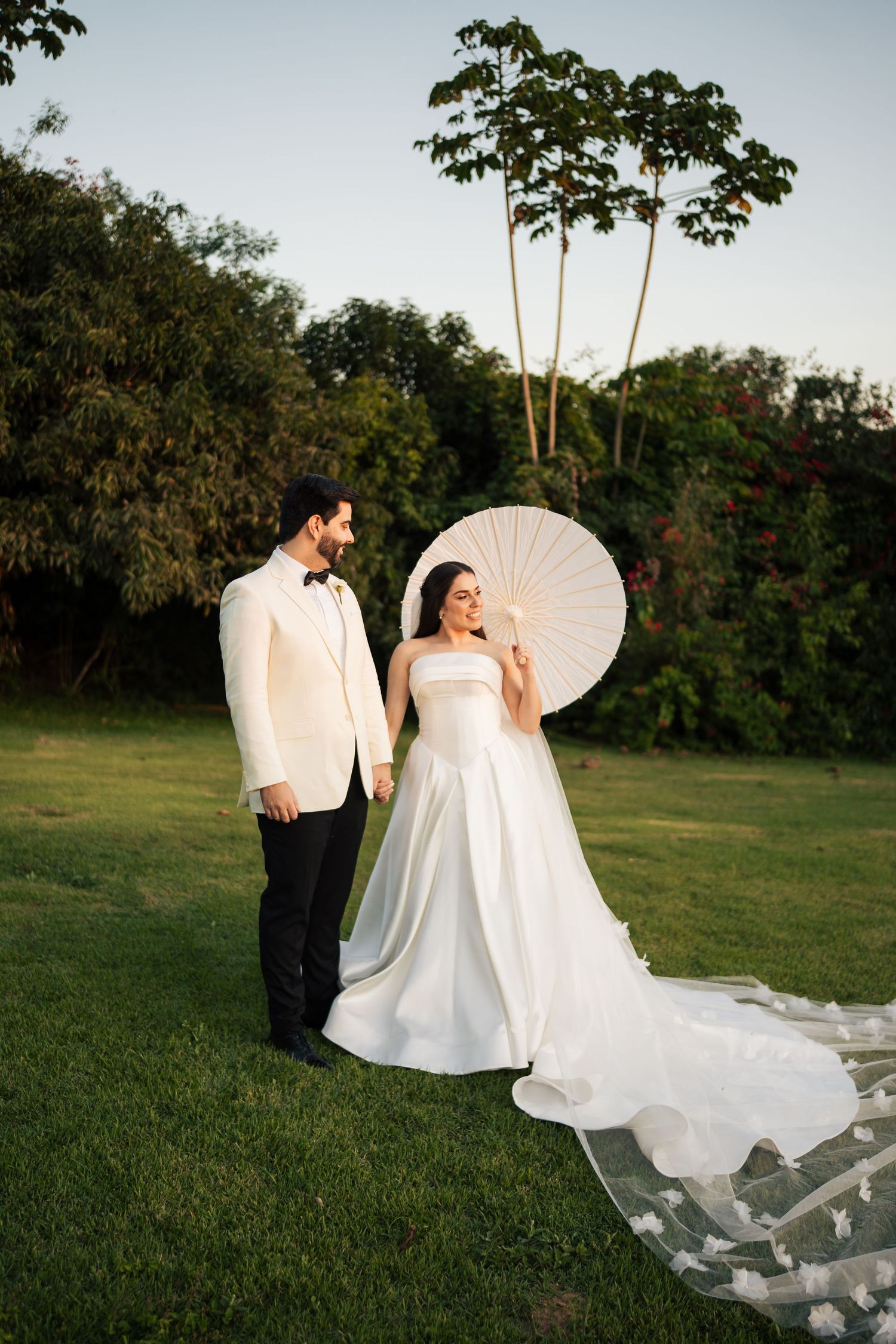 Wedding couple posing with a white parasol in a lush green garden setting at sunset.