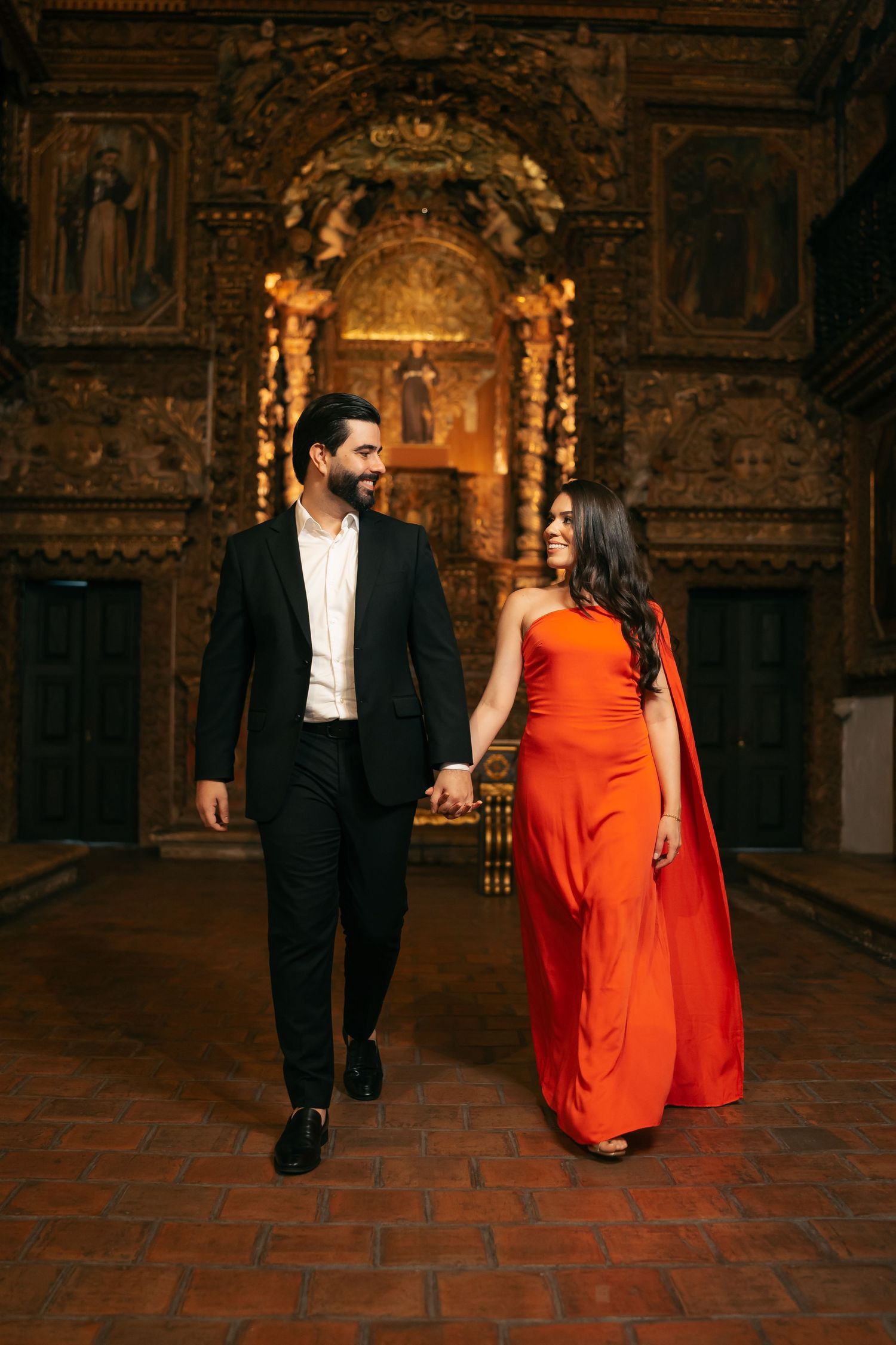 A couple walks through an ornate historic interior with brick floors and golden details.
