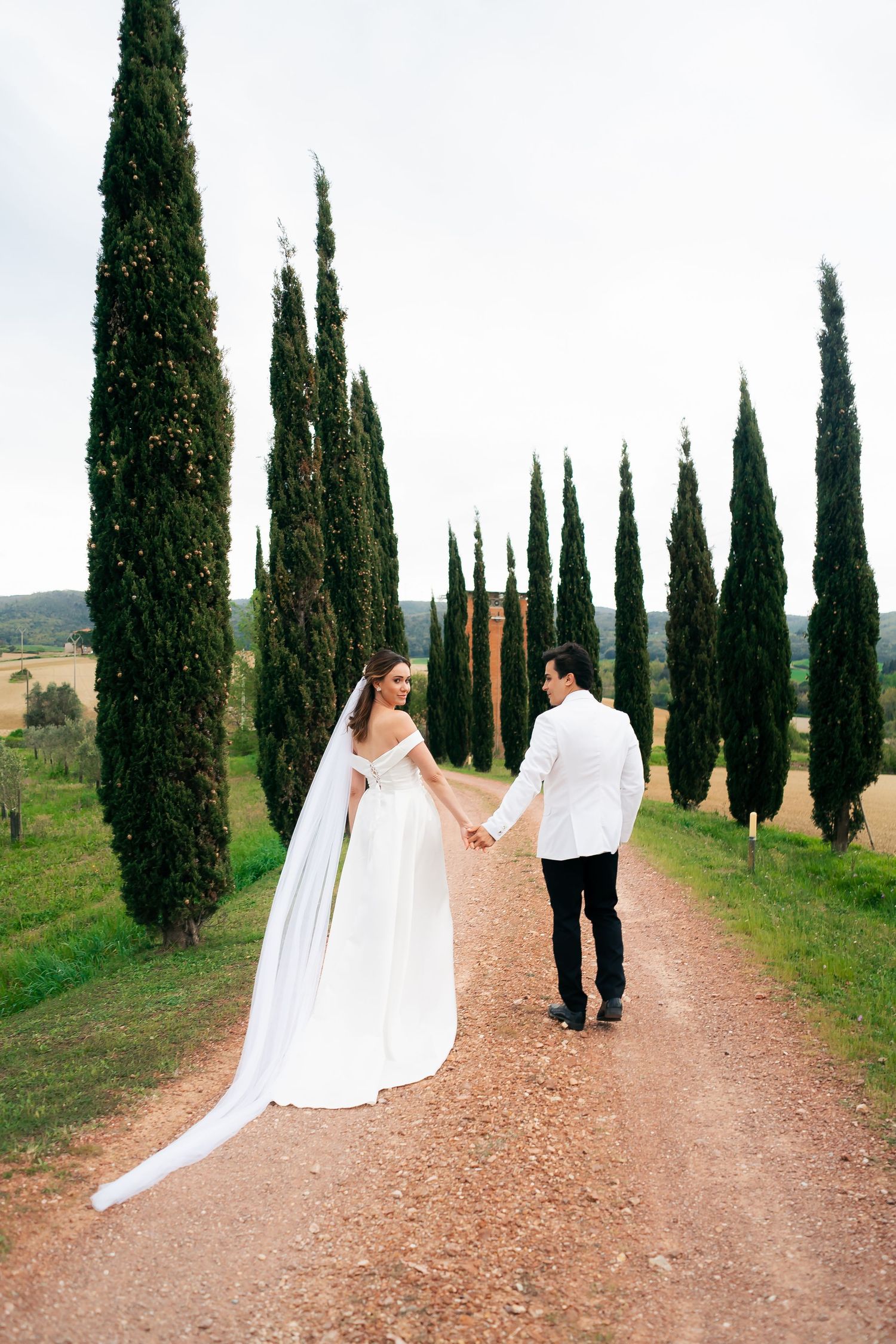 A romantic couple walks down a cypress tree-lined path in the Tuscan countryside on their wedding day.