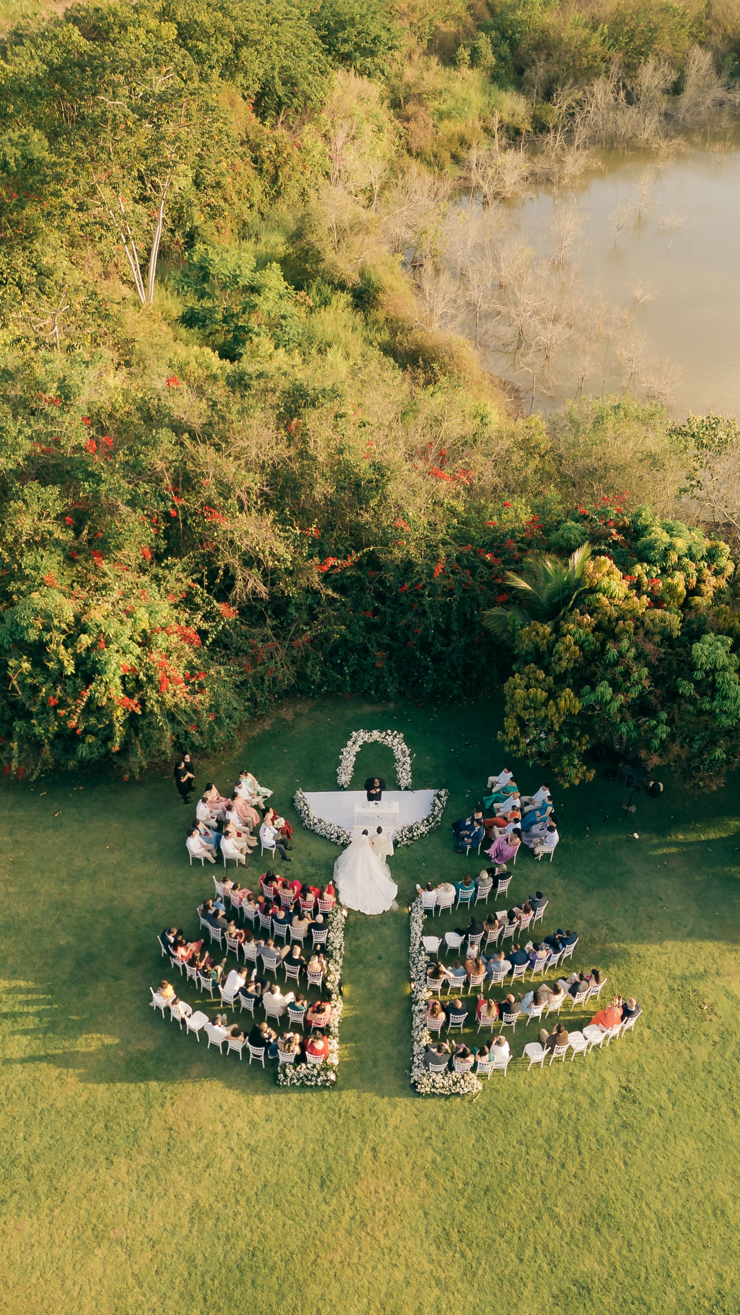 Aerial view of outdoor wedding ceremony with guests arranged in a heart shape on green lawn.