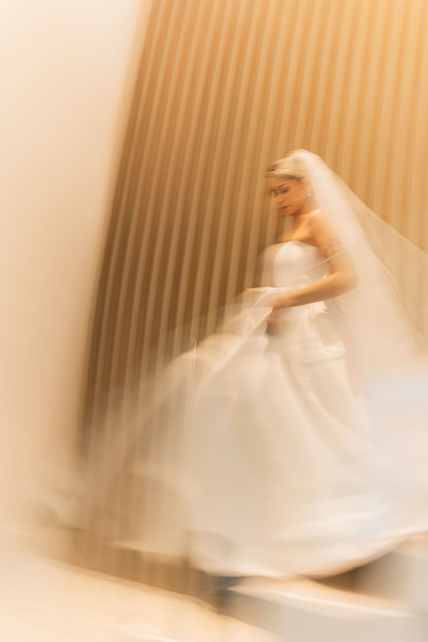 Ethereal motion-blurred image of a bride in a flowing white wedding gown and veil against a warm-toned background.