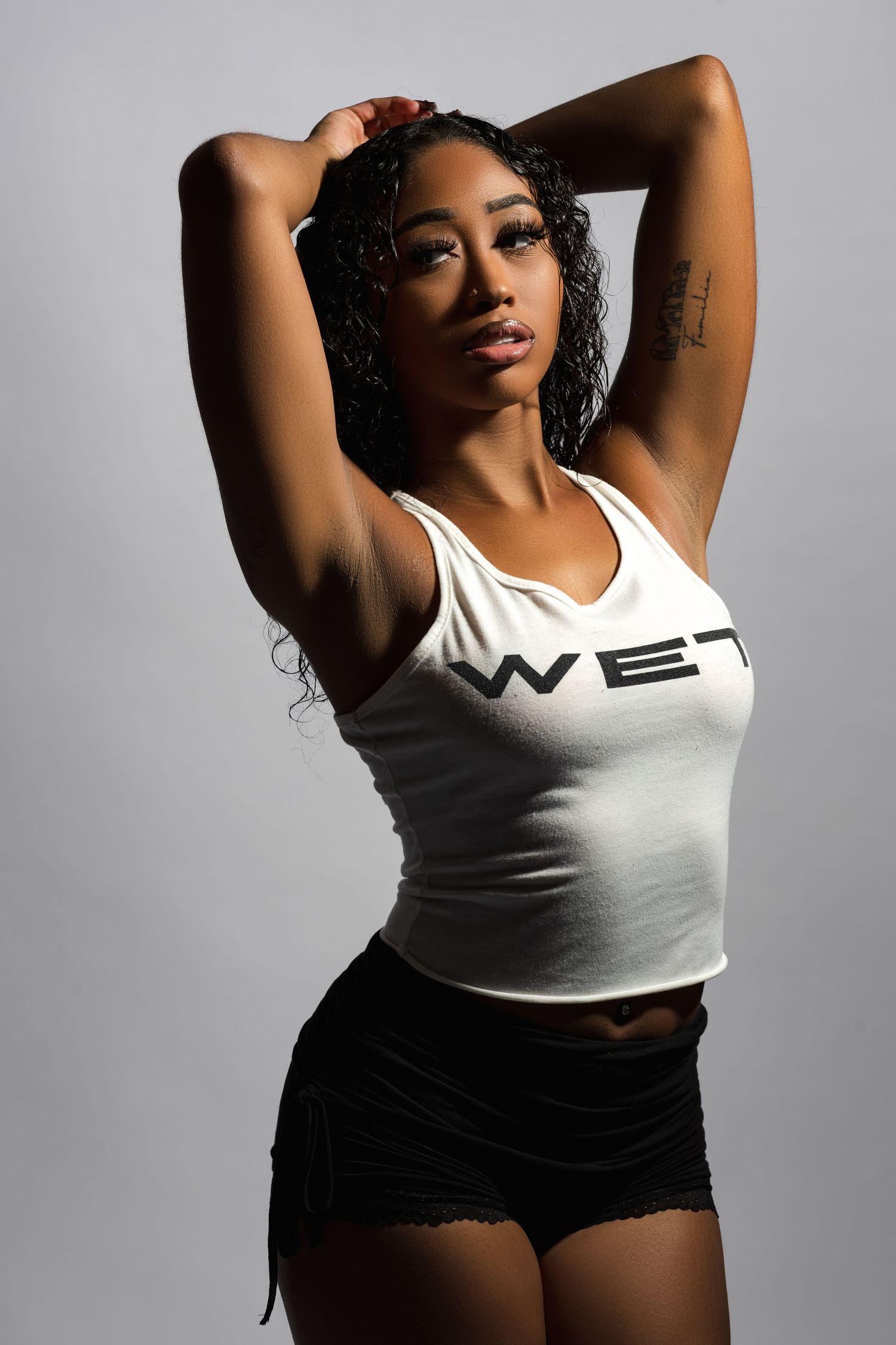 Fashion model poses in white tank top and black shorts against grey studio background.