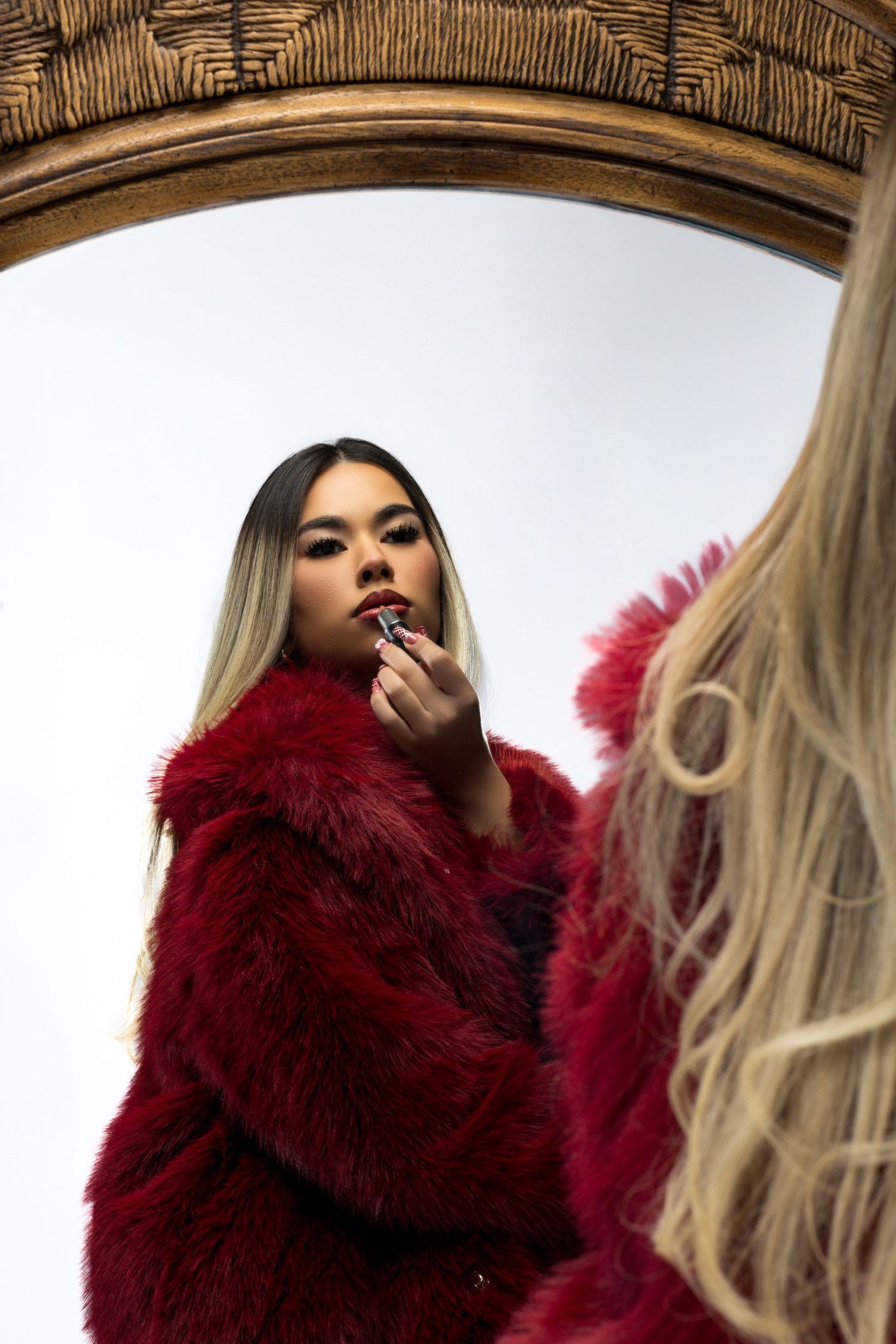 Woman in red fur coat applying lipstick while looking in ornate mirror.