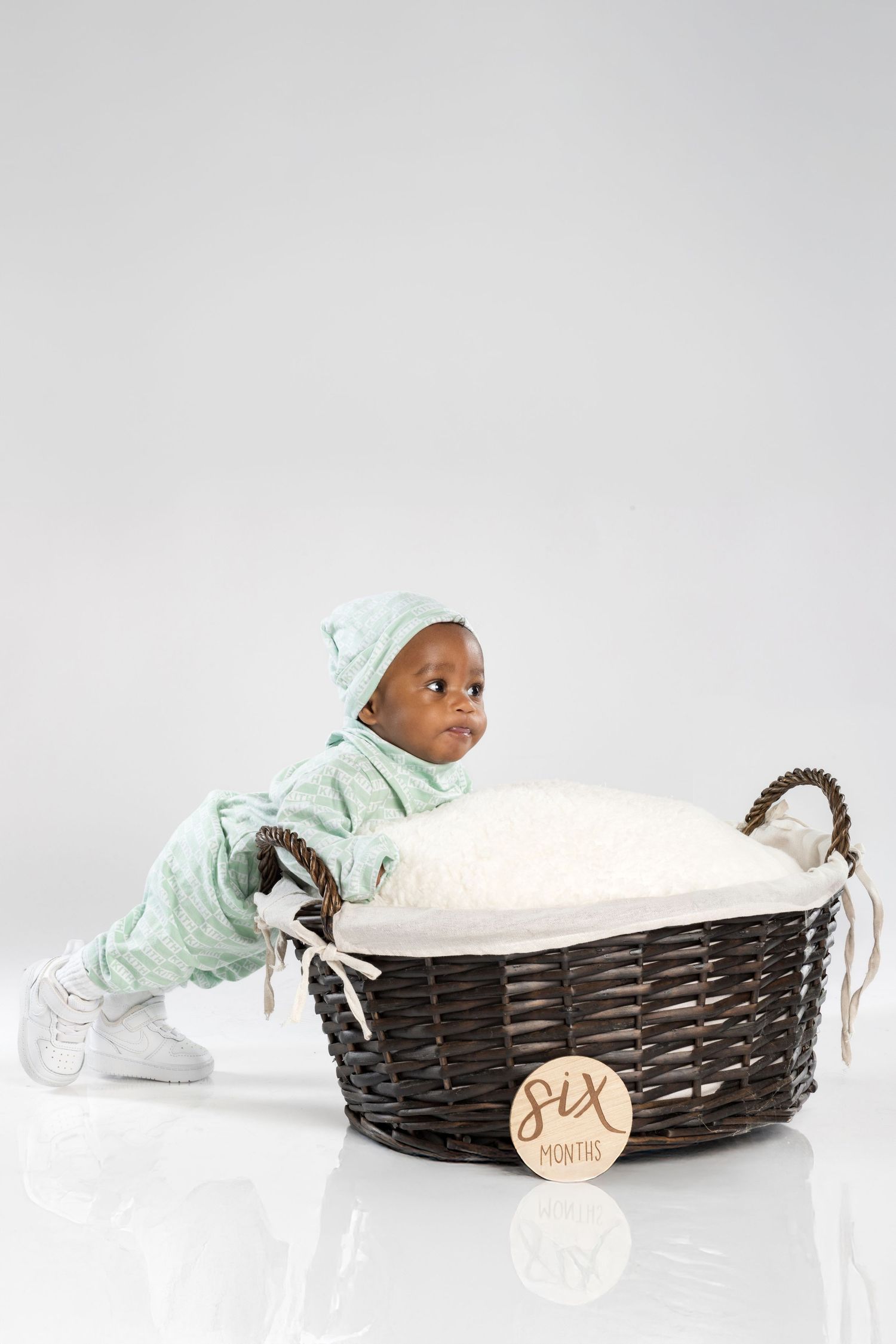 Baby in mint green onesie playfully crawls through dark woven basket during professional photo session.
