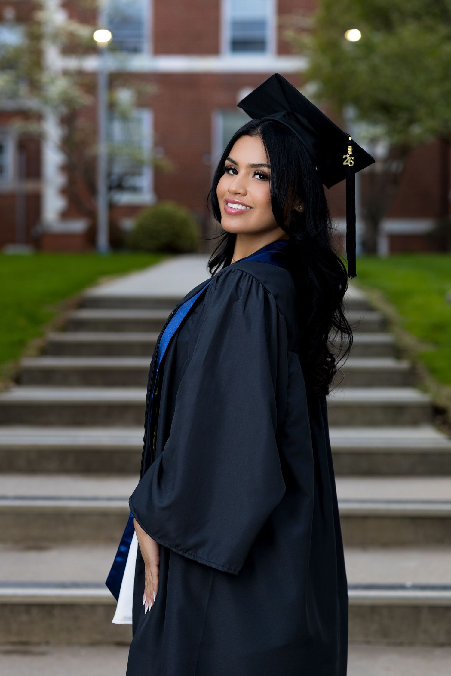 Smiling female graduate in black cap and gown poses outdoors on campus steps holding diploma.