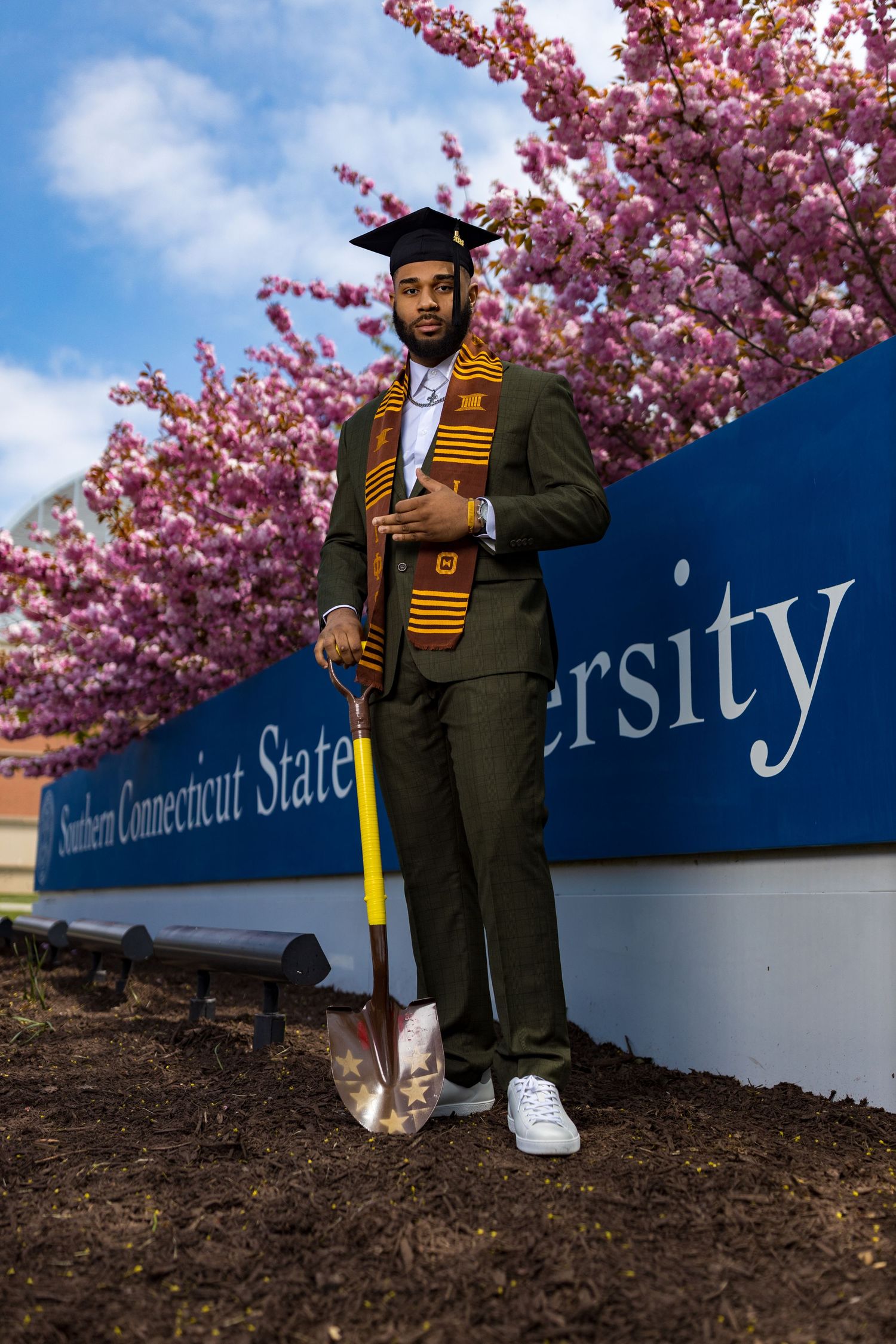 Graduate in traditional cap and gown with kente stole poses near university signage under cherry blossoms.