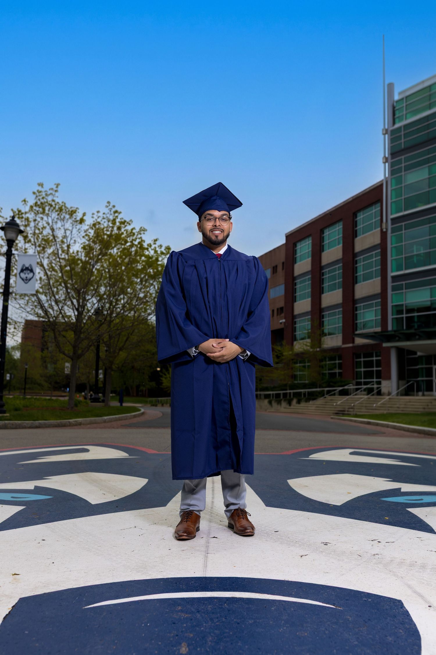 Graduate in navy cap and gown poses proudly on campus roundel with university buildings in background.