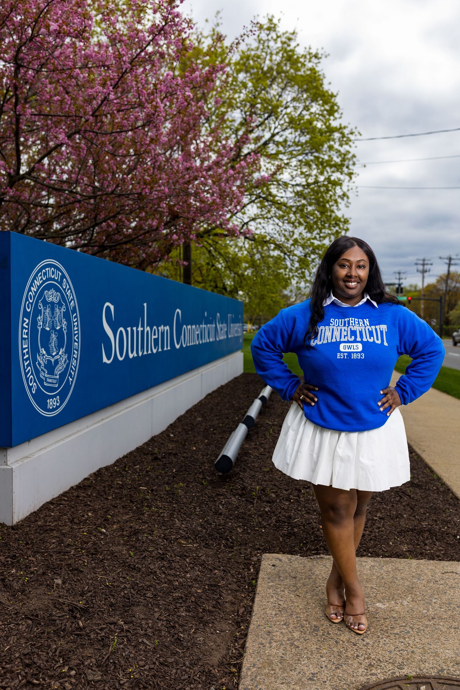 A smiling woman in a blue Southern Connecticut State University sweatshirt poses by the campus entrance sign in spring.