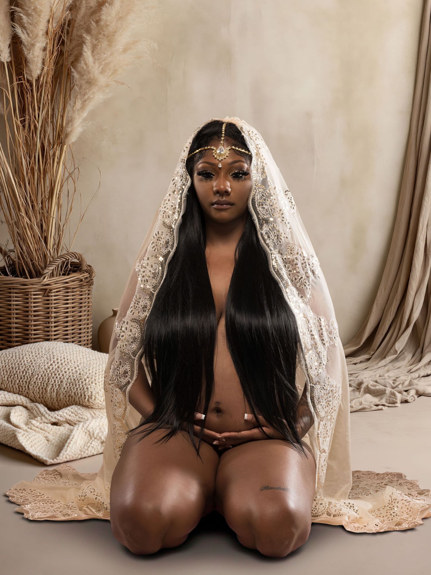 A woman kneels gracefully wearing a lace veil and gold headpiece, with long black hair, in a boho studio setting.
