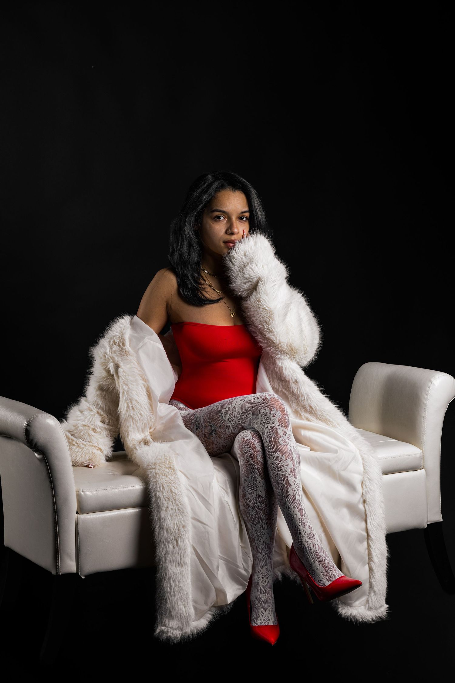 Woman in red bodysuit, white fur coat, lace tights, and red heels sitting on a white chair against black background.
