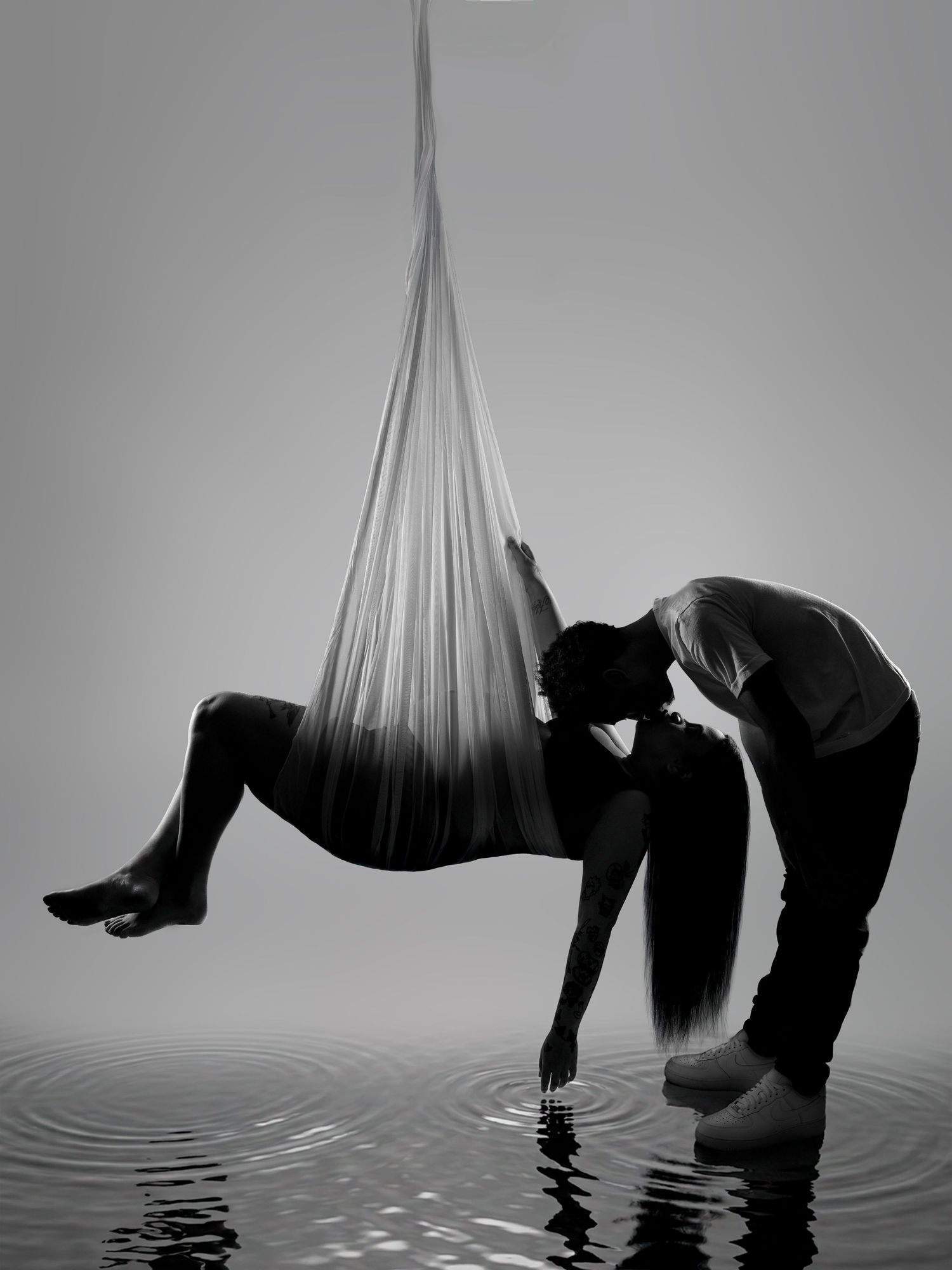 Black and white silhouette of two people sharing a kiss, one suspended in aerial silk over rippling water.