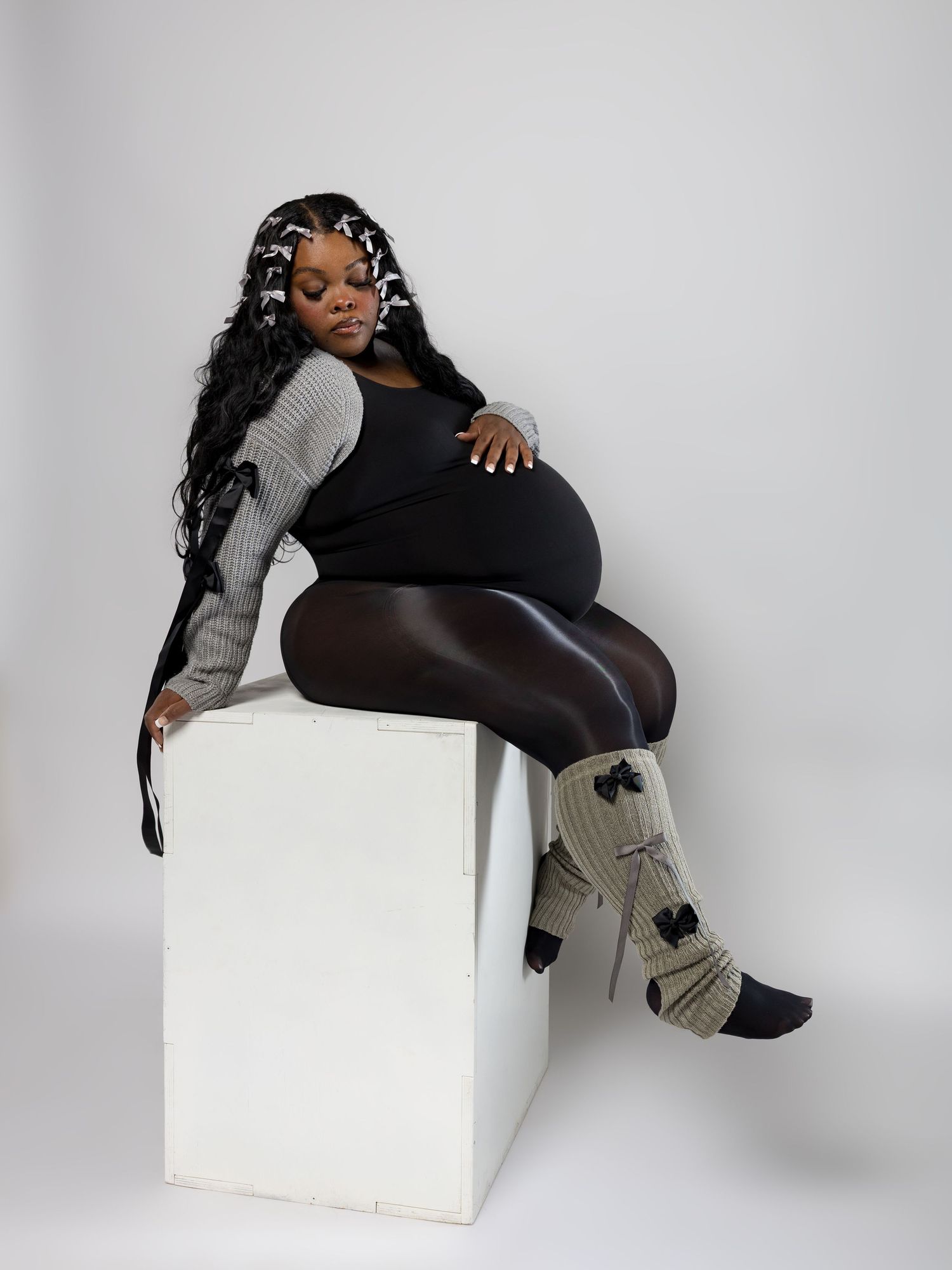 Pregnant Black woman with long braids sits on white cube, wearing black outfit with gray knit leg warmers.