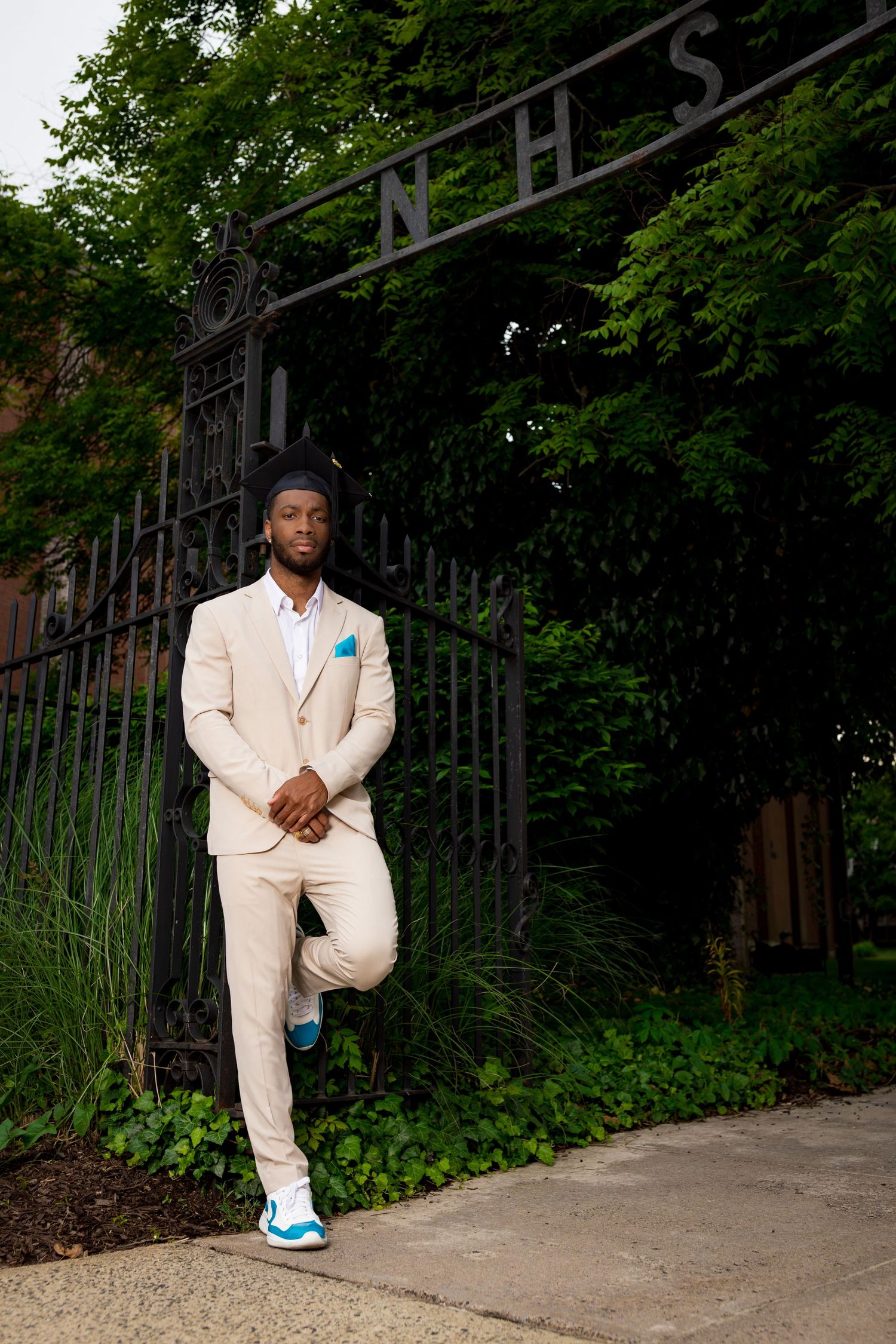 Figure in cream colored suit and blue sneakers posing against ornate iron fence and greenery.