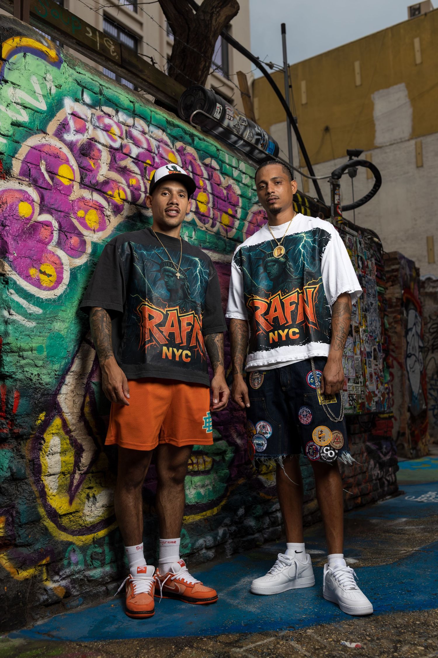 Two people in urban streetwear pose against colorful graffiti wall wearing orange and black outfits.