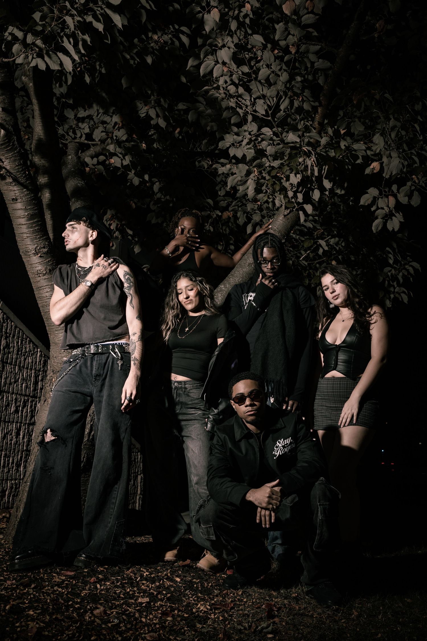 A group of people in dark clothing pose together in a moody outdoor nighttime photoshoot against trees and foliage.