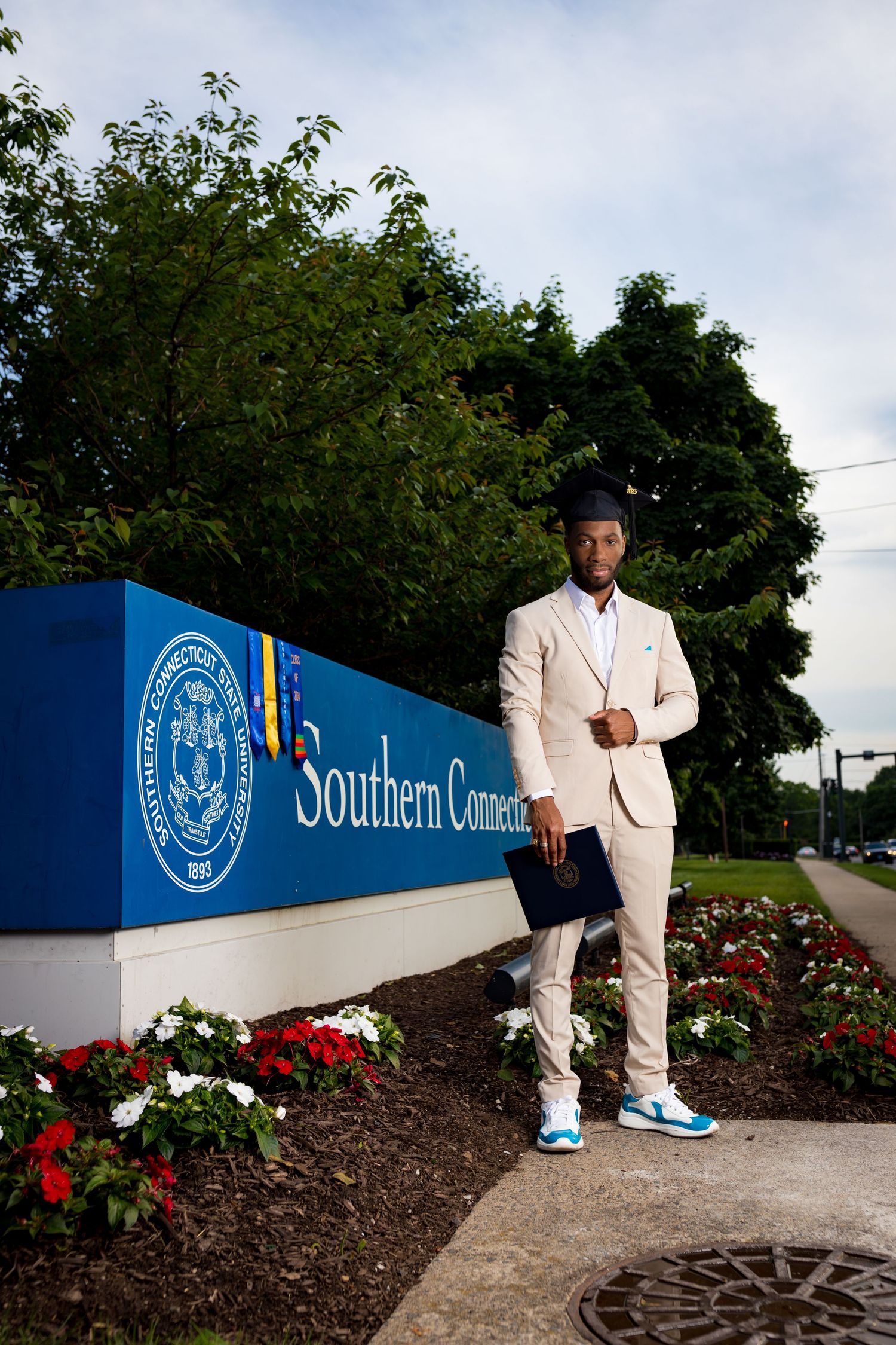 Person in light suit poses near Southern Connecticut university entrance sign with red flowers.