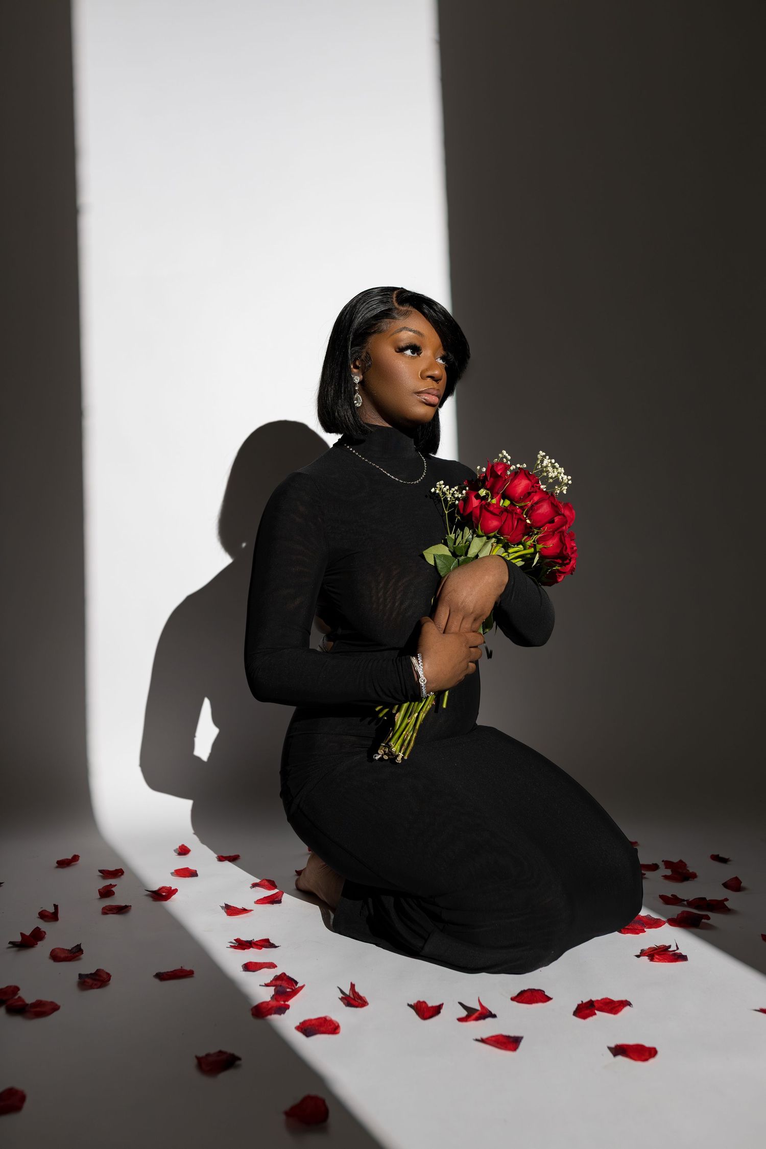 A figure in a black outfit sits on white floor with scattered red rose petals in a dramatic studio lighting setup.
