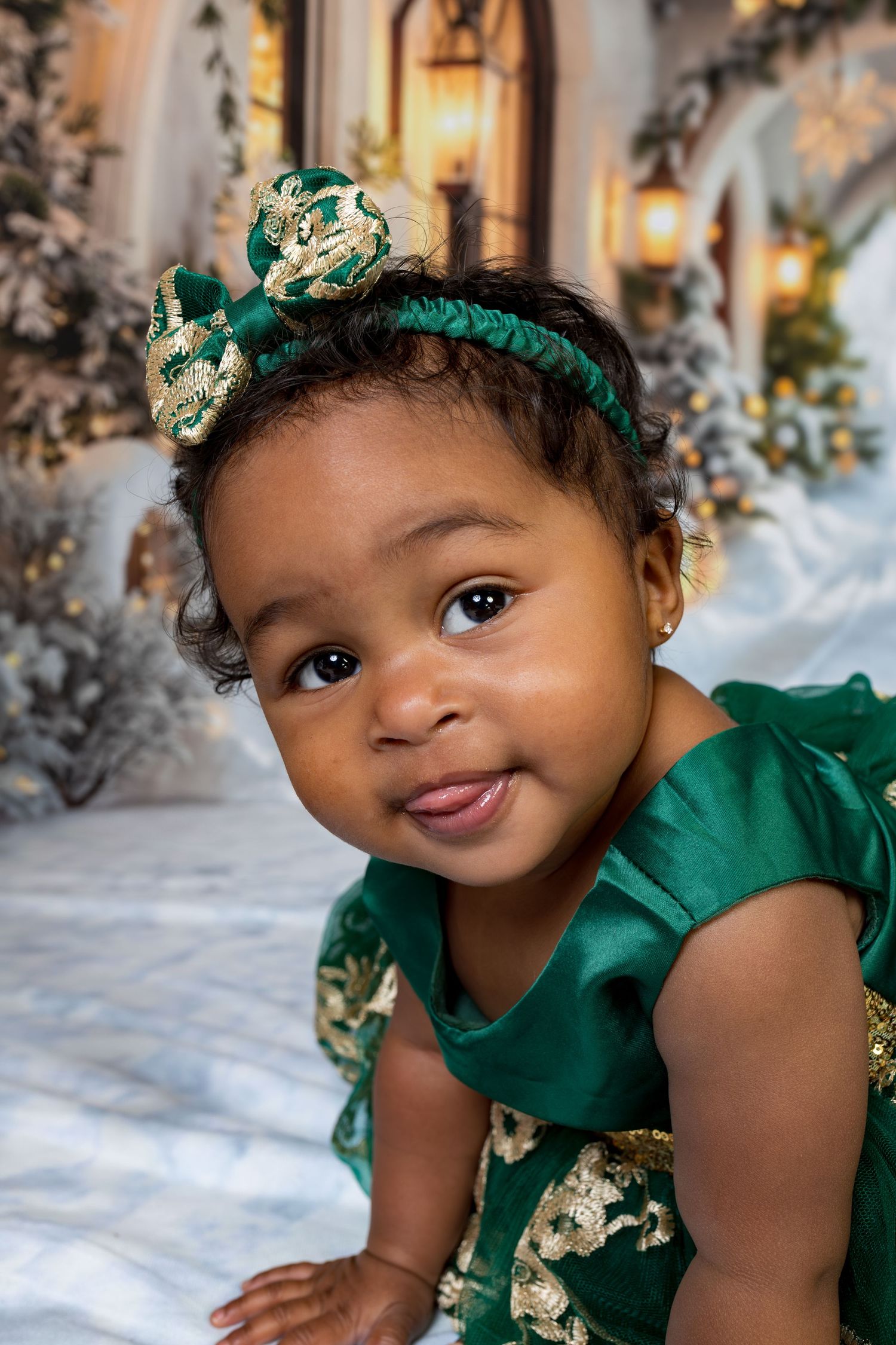 Baby wearing emerald green dress and floral crown smiles while crawling on white backdrop with winter decor.
