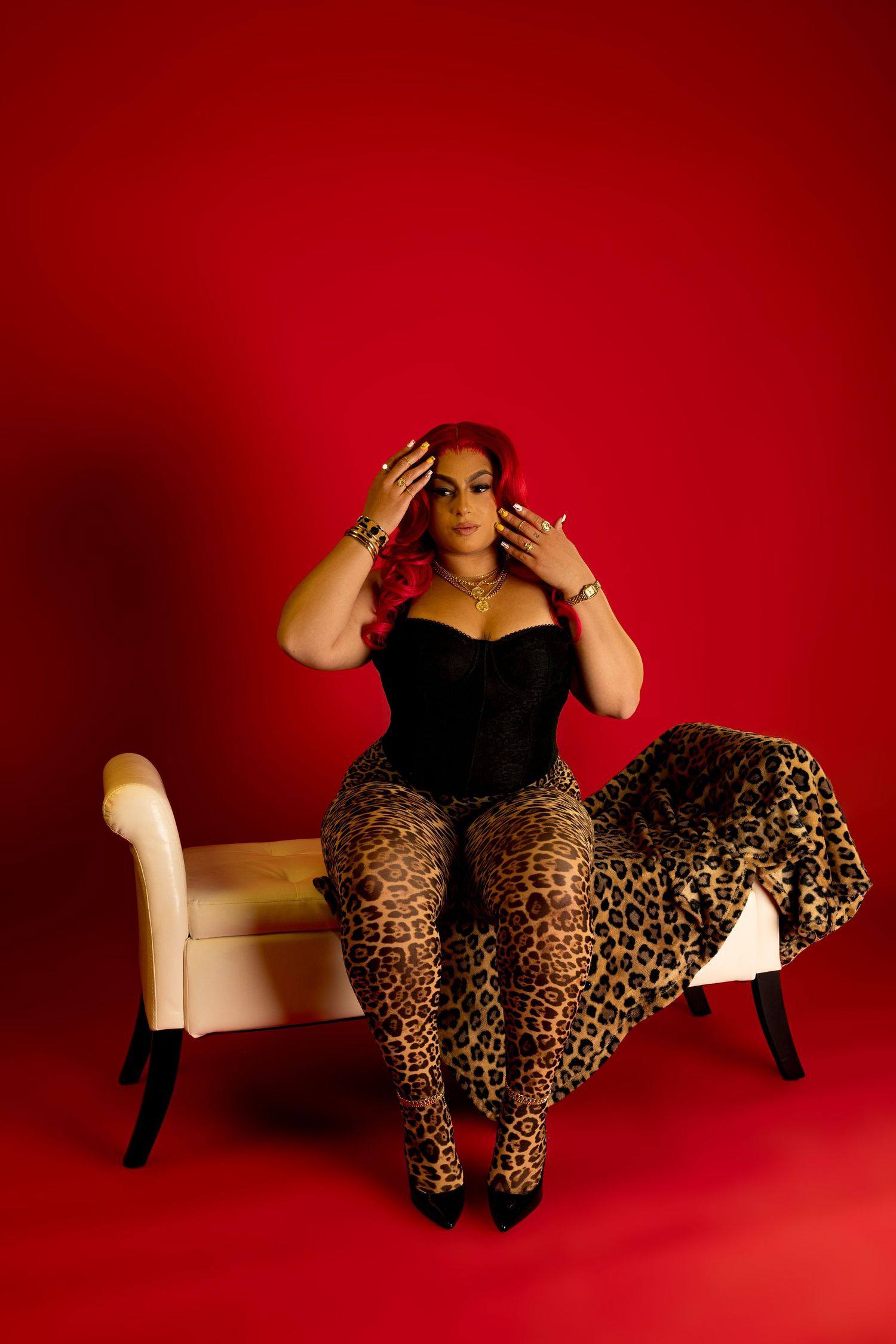 Woman in black corset and leopard print pants sits on a chaise lounge against a bold red background.