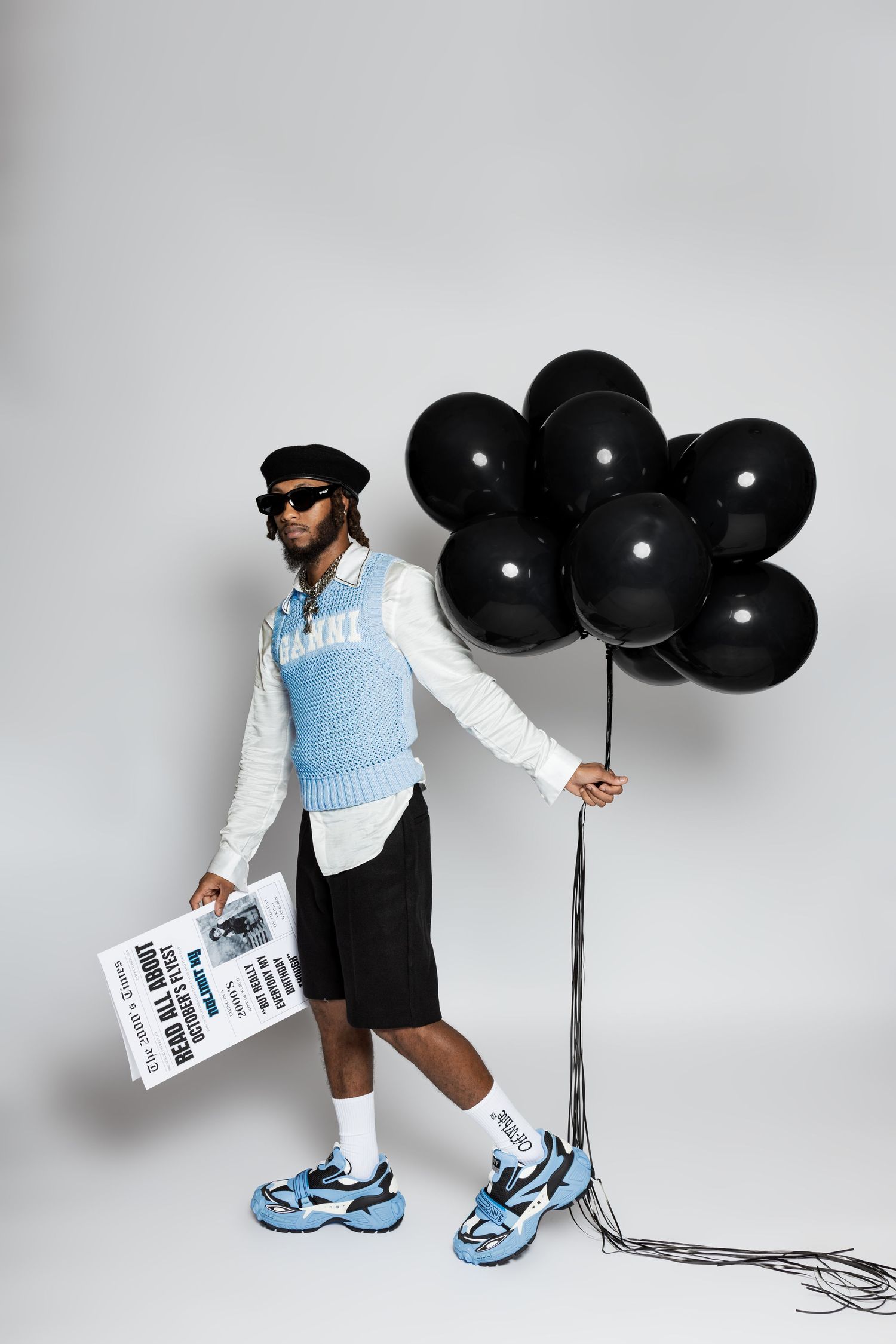Stylish figure in blue and white outfit holding black balloons and a newspaper against white backdrop.