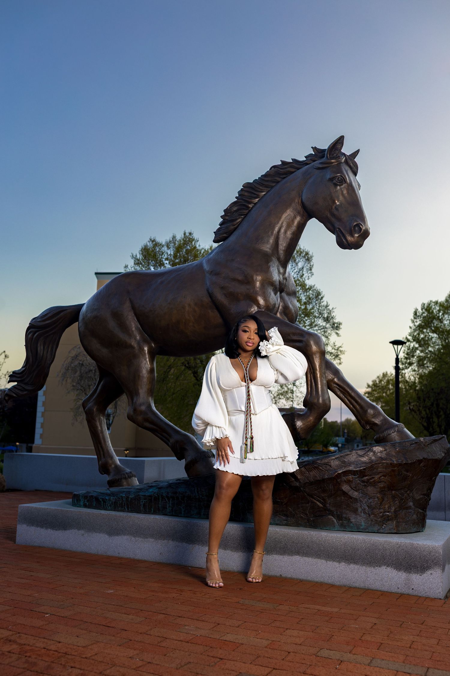 Woman in white dress poses before a bronze rearing horse statue on brick plaza at dusk.