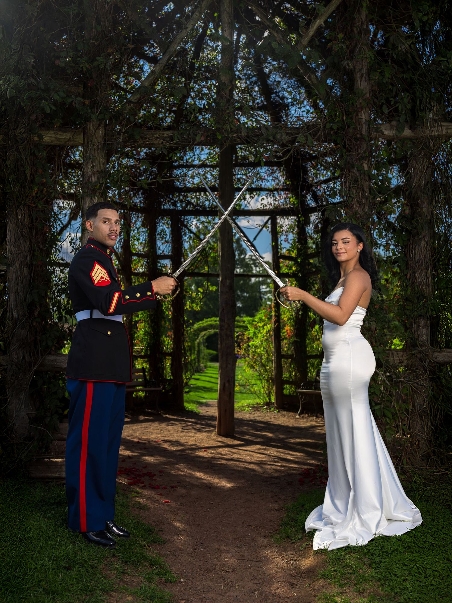 Couple in formal attire poses playfully under garden trellis during outdoor photoshoot.