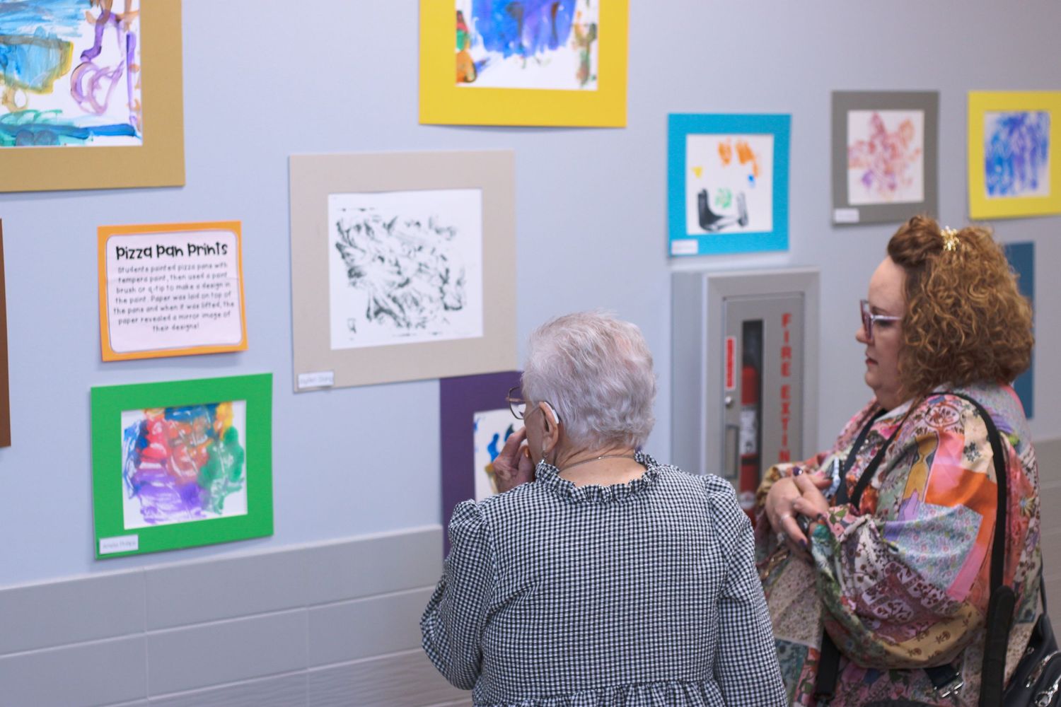 Art gallery visitors admire colorful artwork displayed on gray walls in a well-lit exhibition space.