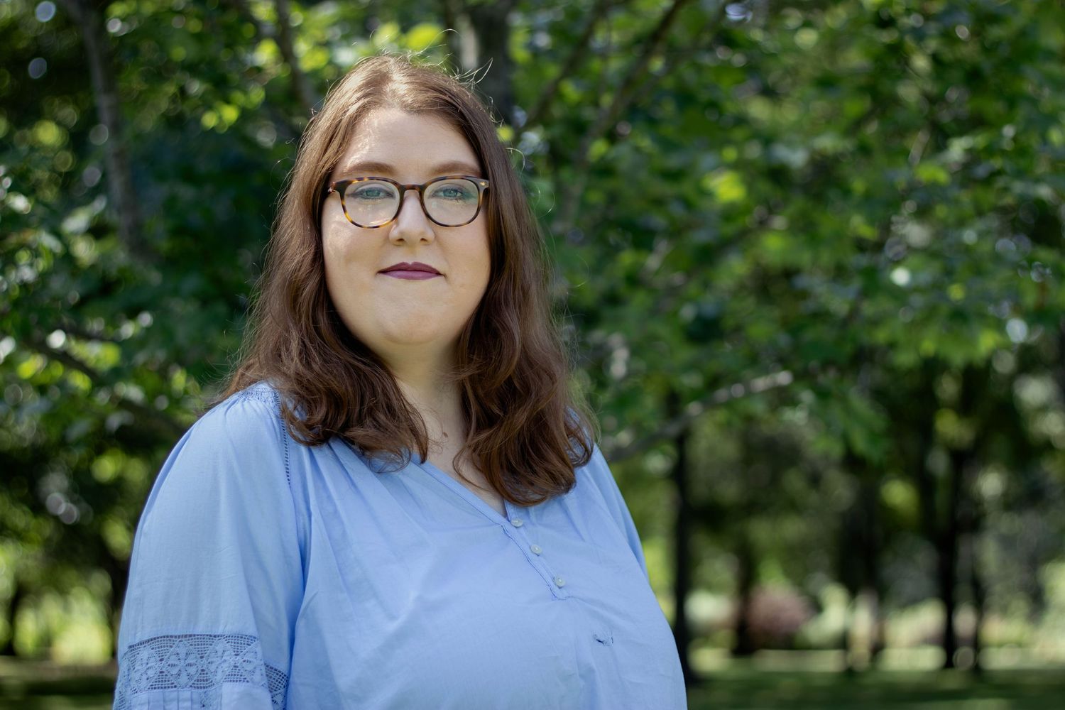 Woman with long brown hair and round glasses wearing a light blue blouse stands outdoors in a sunlit park surrounded by green trees.
