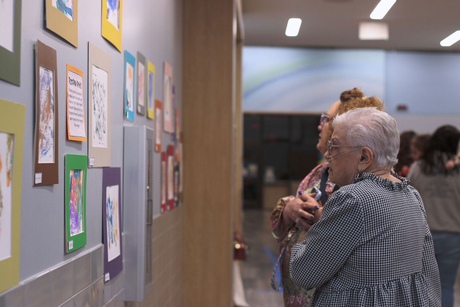 Art gallery visitors admire colorful artwork displayed on gray walls in a well-lit exhibition space.