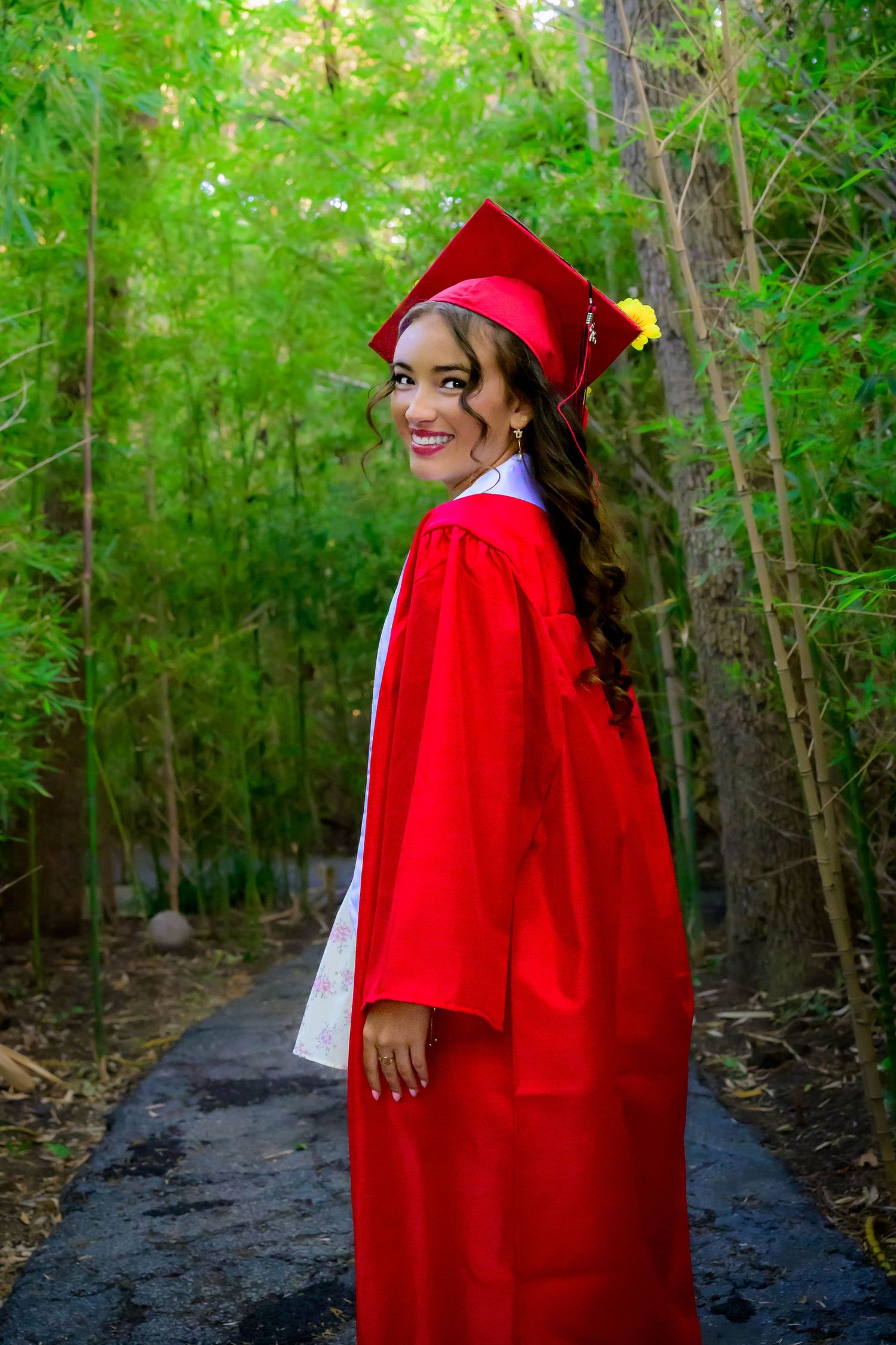Smiling graduate in red cap and gown poses on a tree-lined path during outdoor graduation photo session.