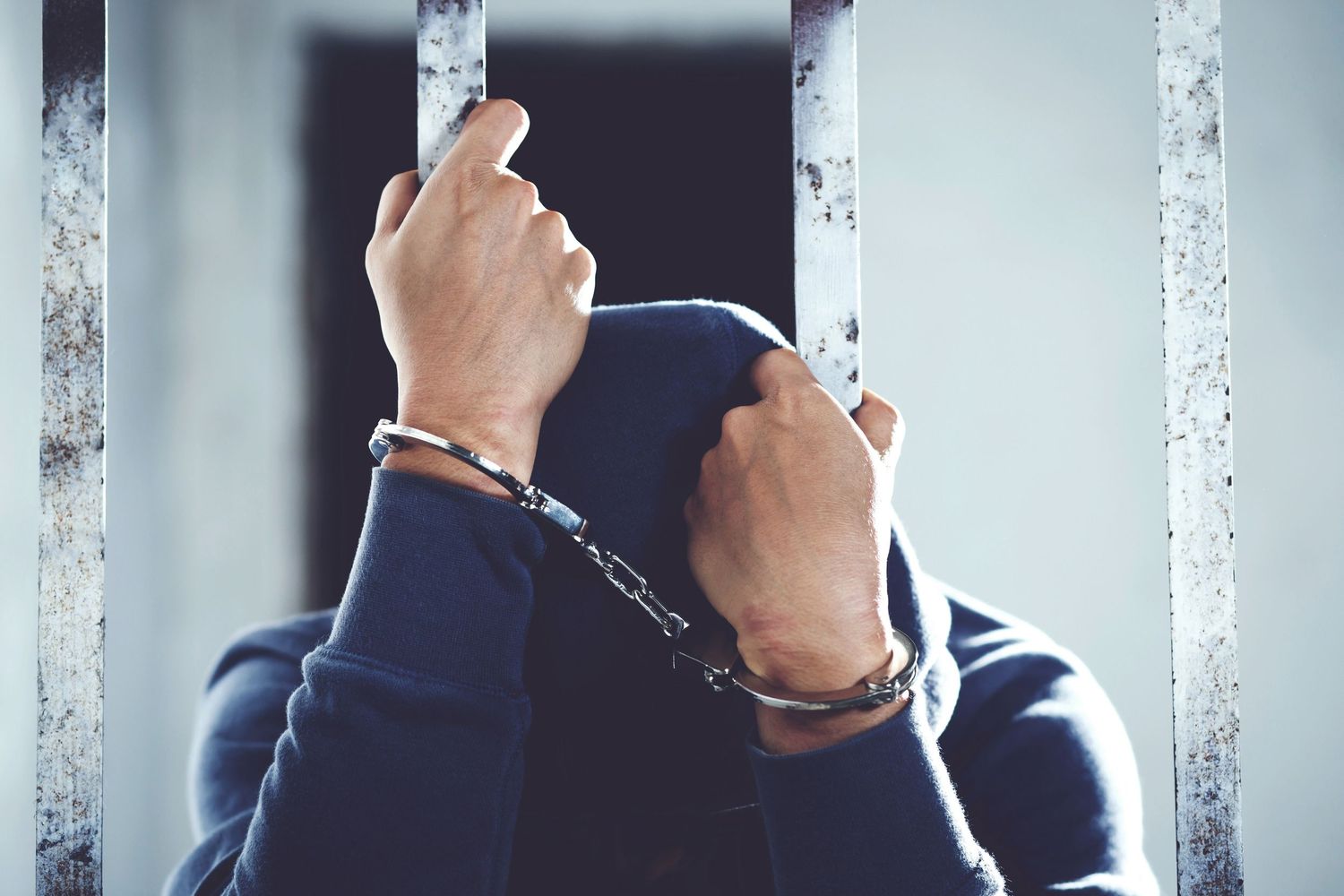 Close up view of hands in handcuffs gripping metal bars through prison cell window.