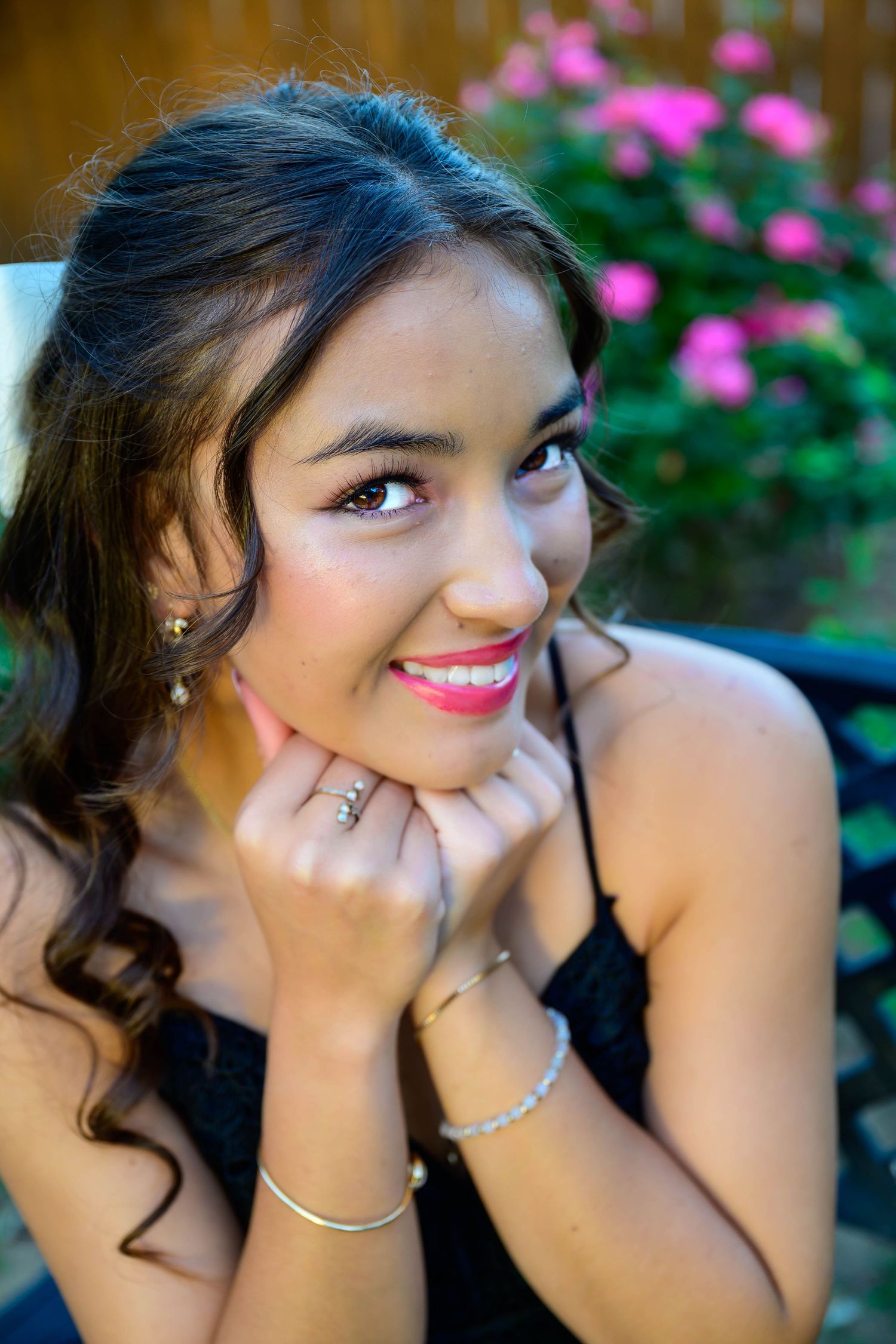 Smiling young woman with long wavy dark hair rests her chin on her hands, wearing a black dress and silver bracelets near pink roses.